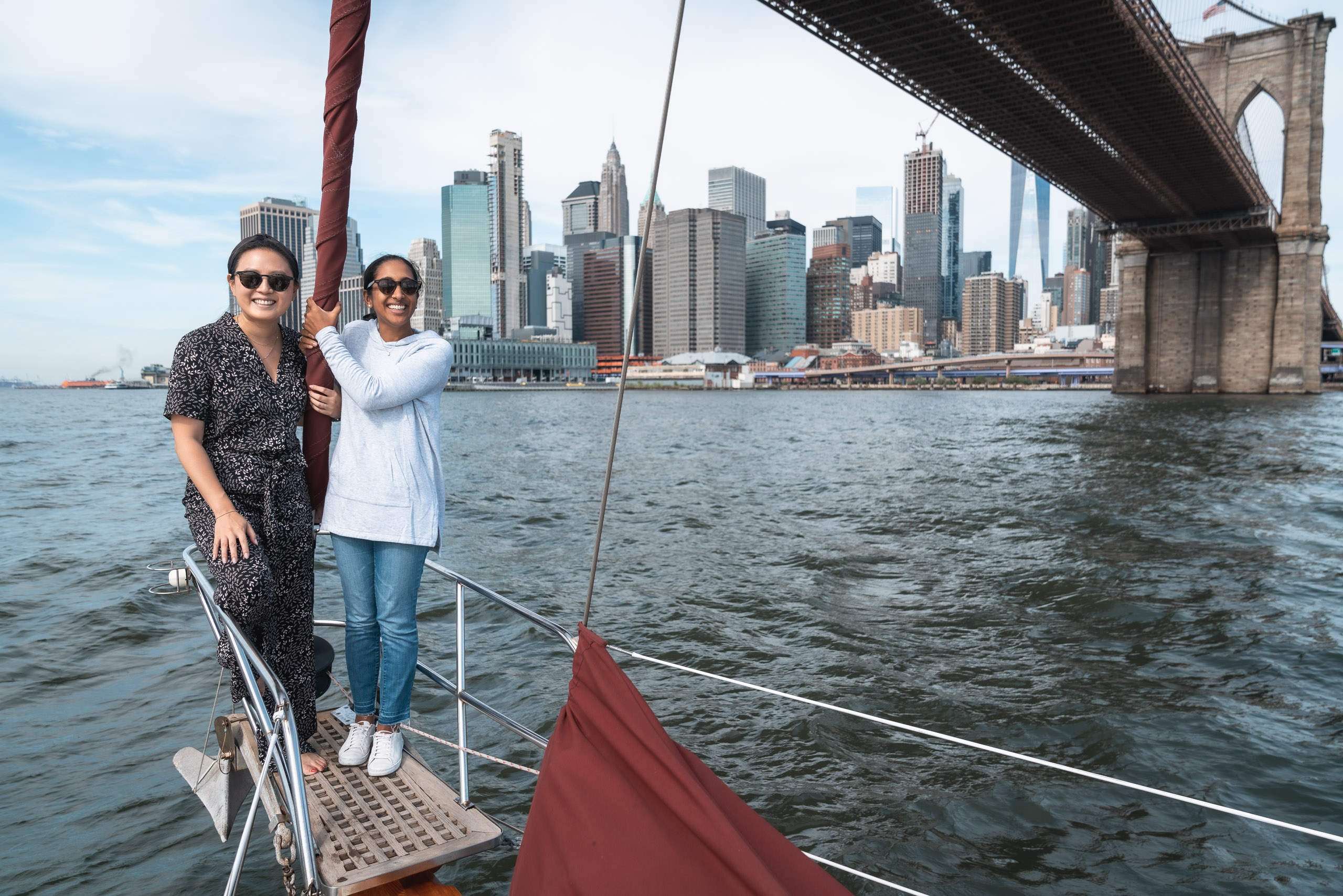 Two women stand on a sailboat's deck beneath the Brooklyn Bridge, with Manhattan's skyscraper-lined skyline visible across calm harbor waters under partly cloudy daylight