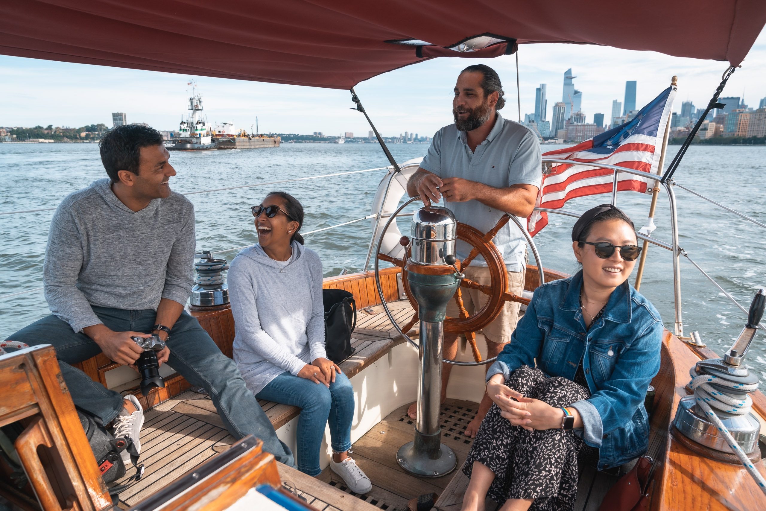 Two people operate a wooden sailboat on calm water with an American flag flying, while the Manhattan skyline stretches across the distant shoreline under clear skies