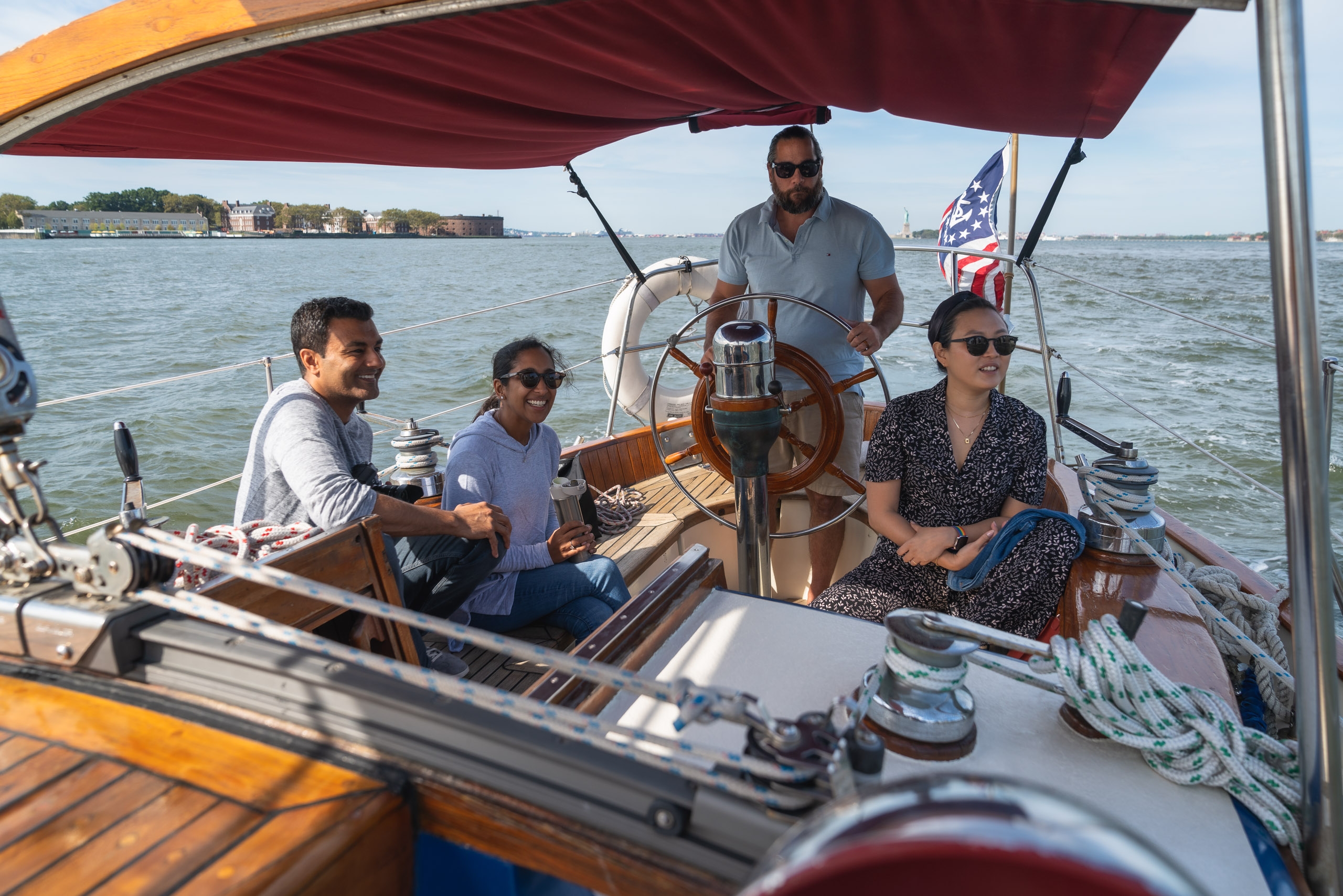 Four people sail on a wooden boat beneath a burgundy canvas awning, with one man at the helm and an American flag visible against calm harbor waters and distant shoreline