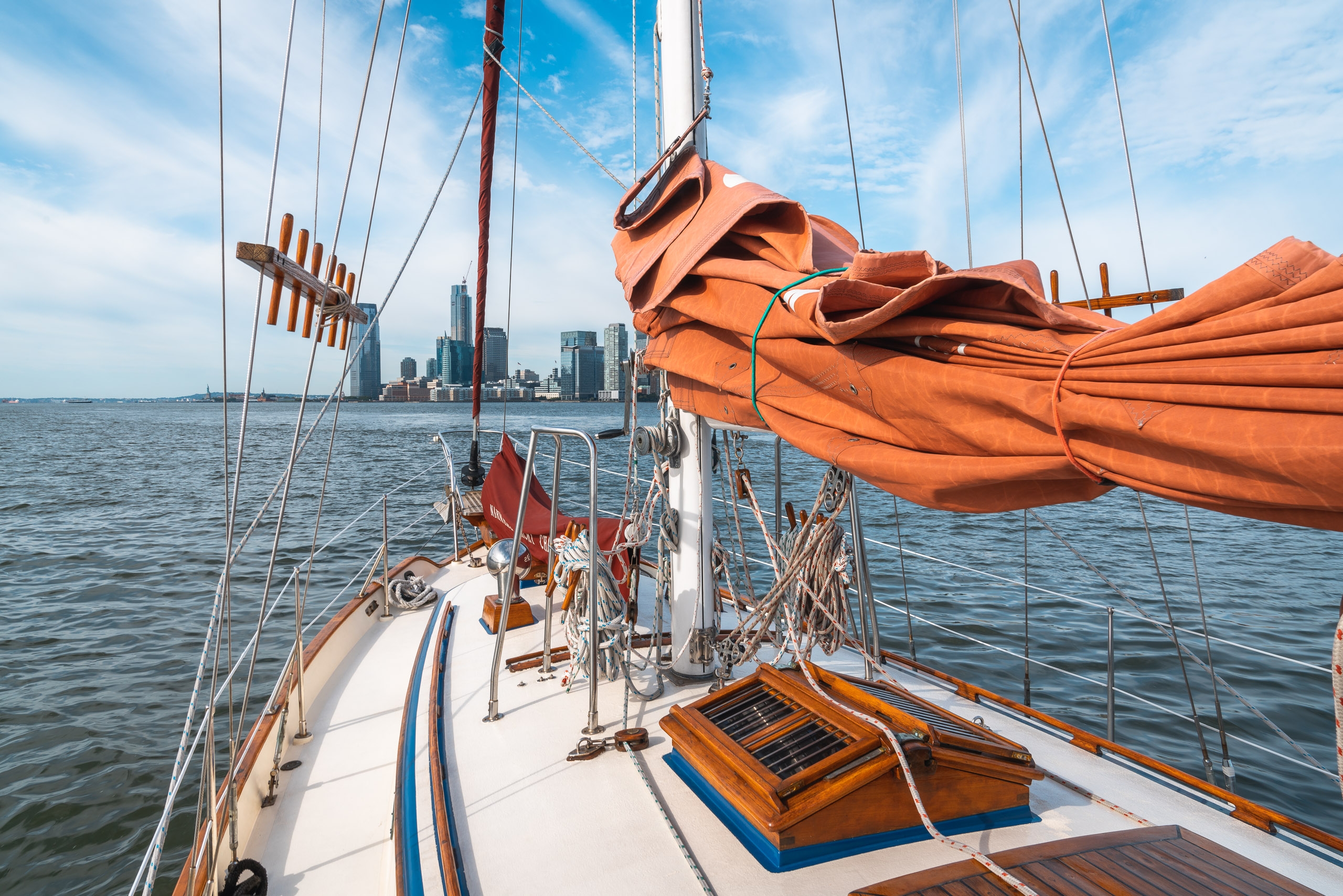 A traditional sailing vessel with furled orange sails and rigging sits on calm harbor waters, with the Manhattan skyline visible across the bay under clear blue skies