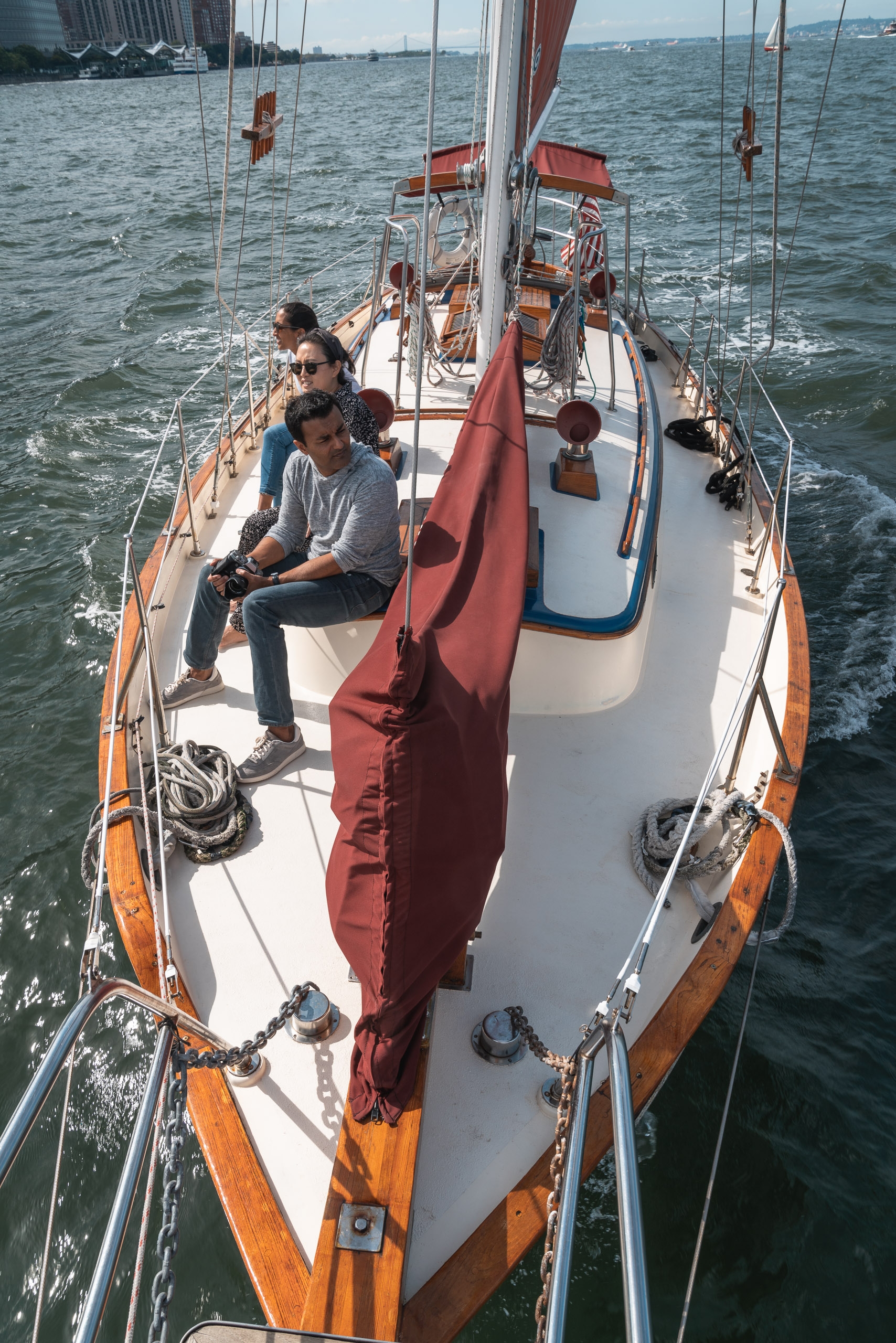 A sailing yacht with white and burgundy canvas moves through choppy green water, carrying several passengers seated along the wooden-trimmed deck under clear skies.