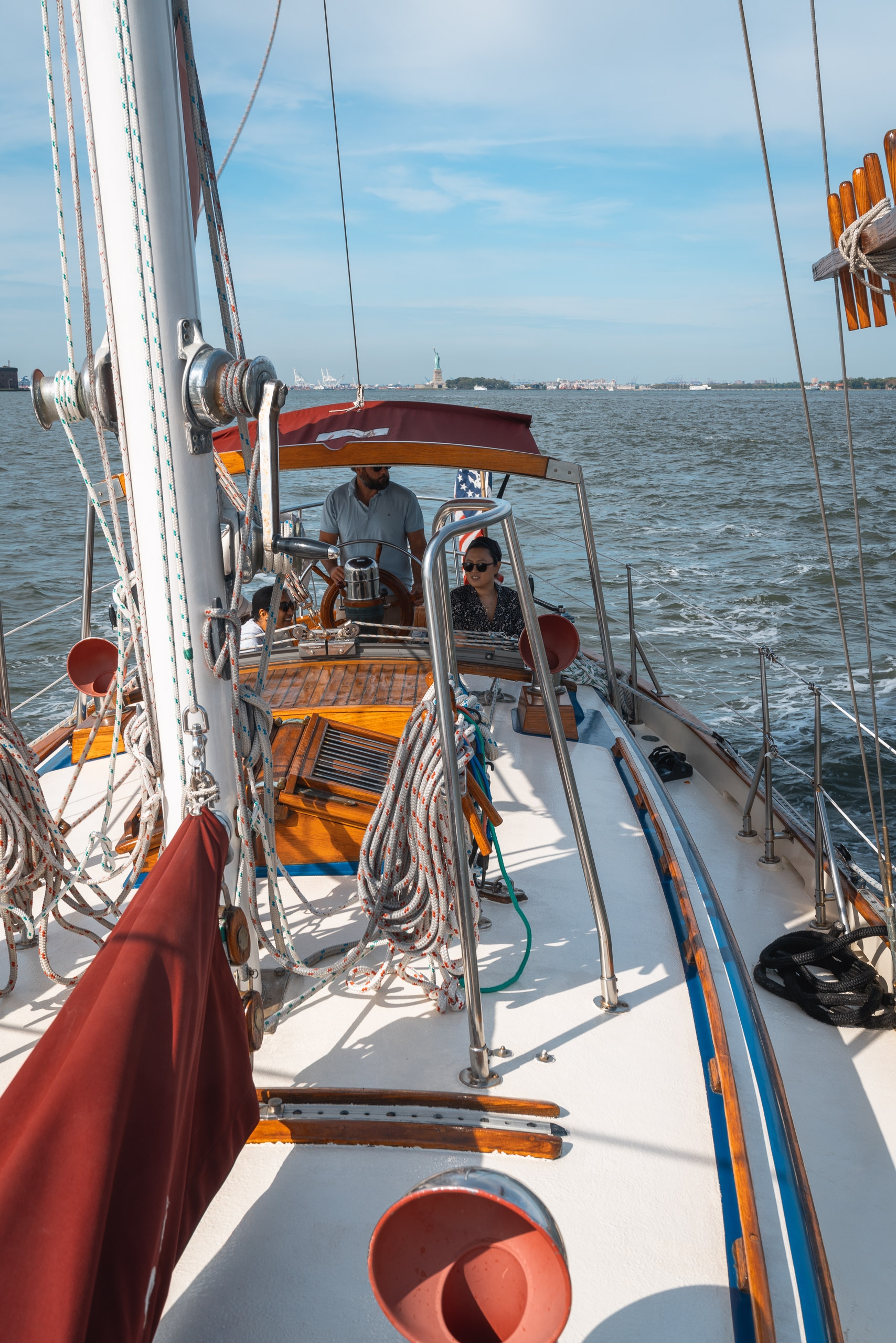 Sailors operate a wooden-trimmed sailboat under full canvas, with rigging visible in foreground and a distant coastline beneath clear blue sky