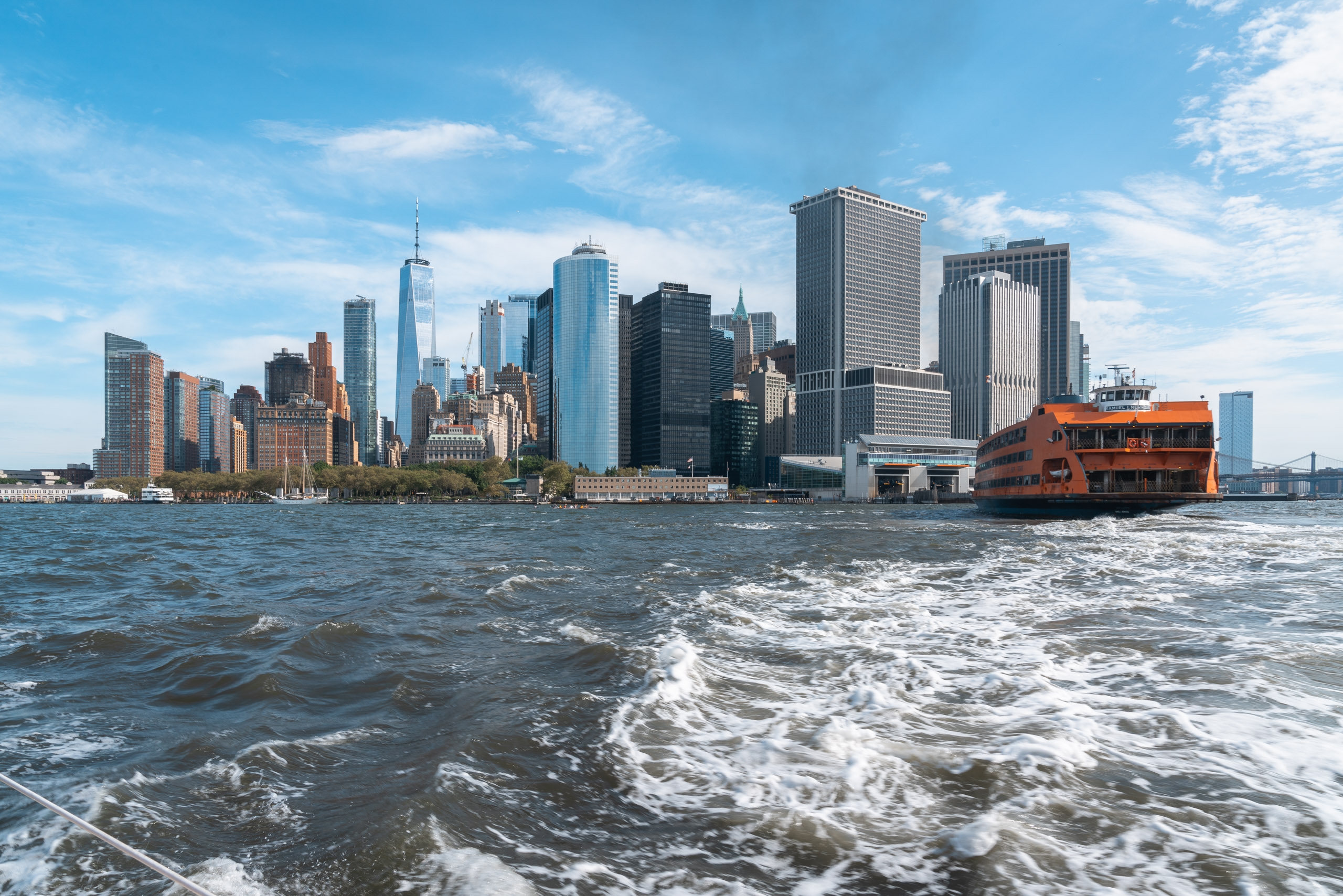 Manhattan's downtown skyline rises beyond choppy harbor waters, with an orange Staten Island Ferry passing in the foreground under clear blue skies