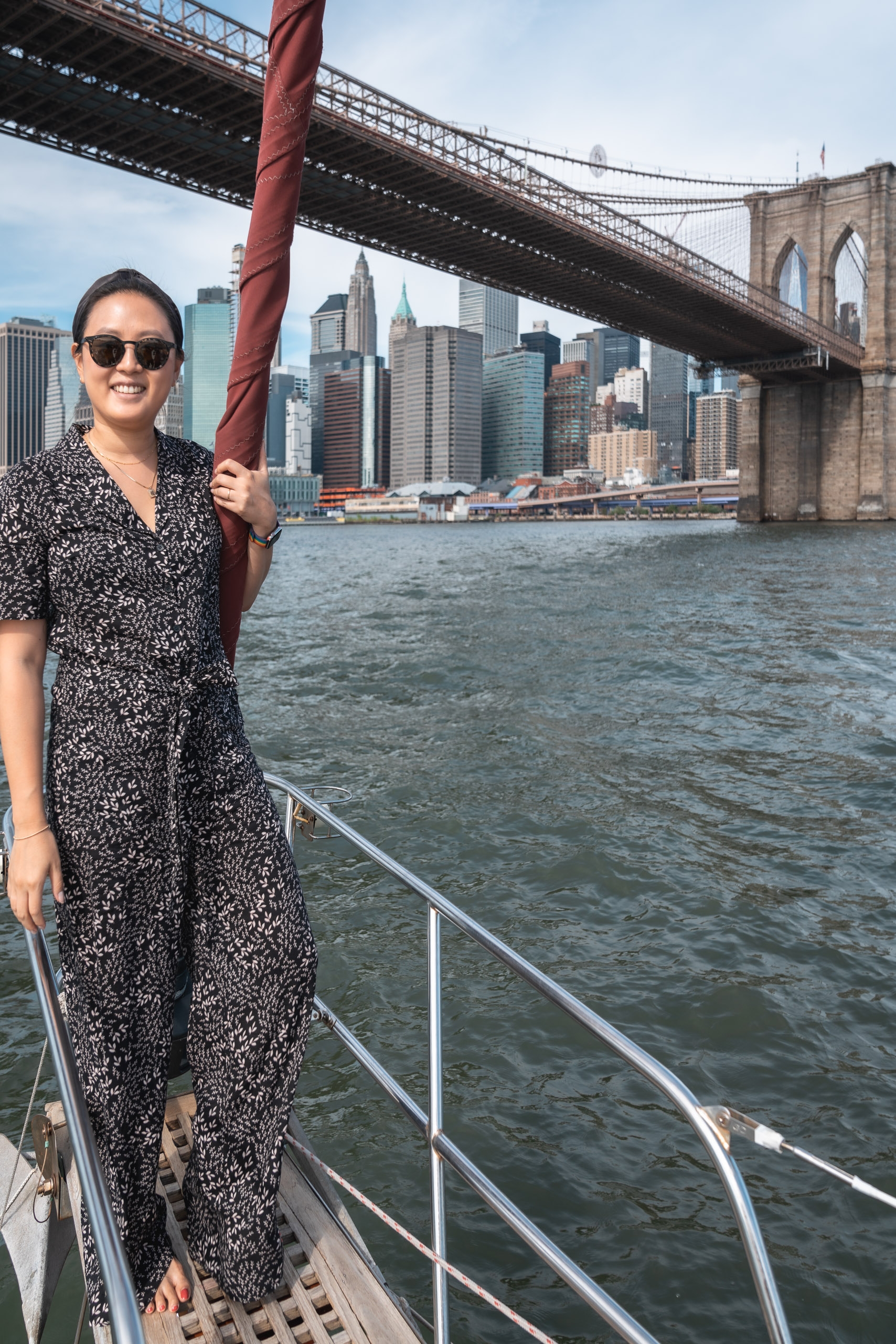 A woman in sunglasses and a patterned black dress stands on a boat deck, holding a red cable beneath the Brooklyn Bridge with Manhattan's skyline visible across the East River