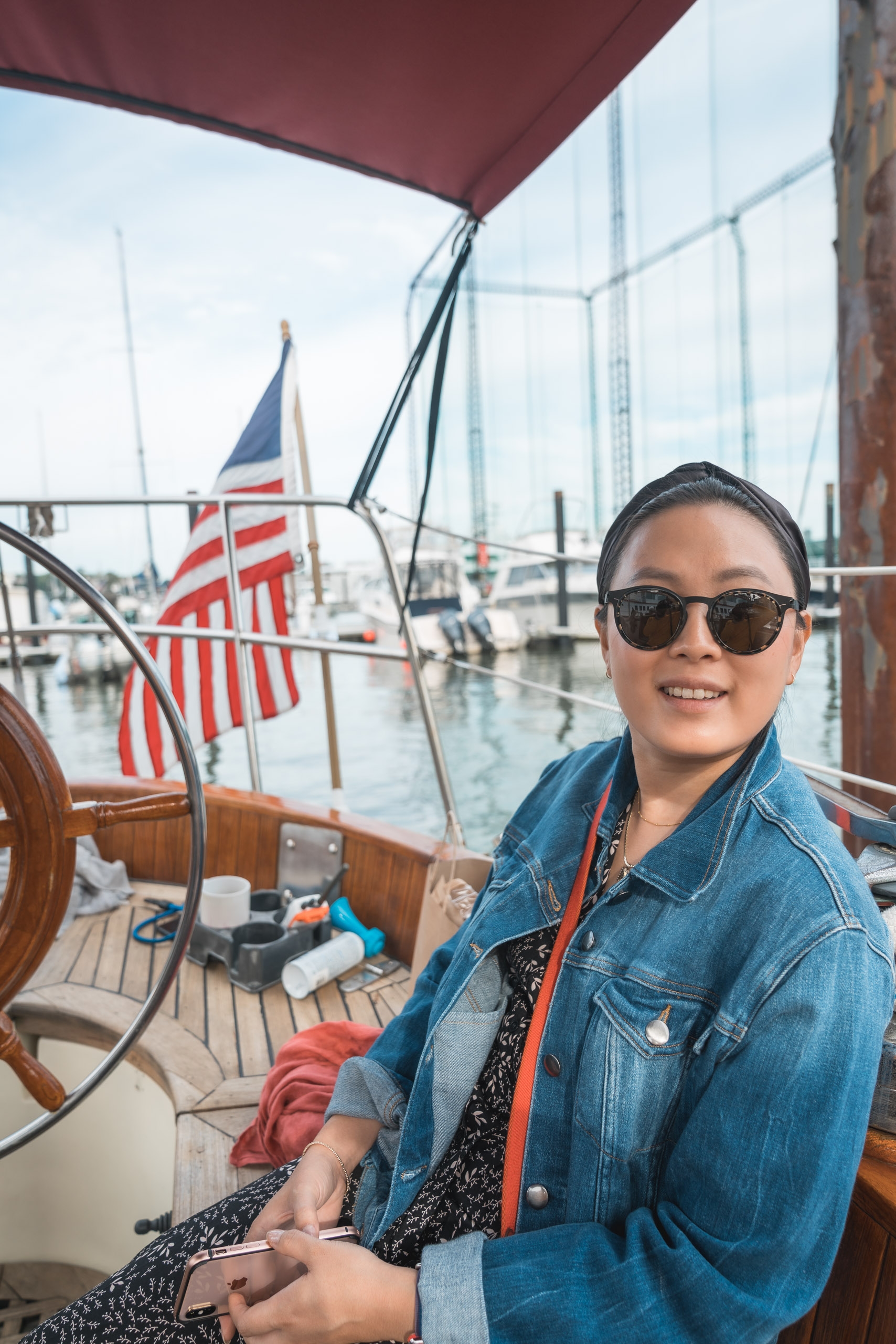 A woman in sunglasses and denim jacket sits on a sailboat deck under a burgundy awning, with American flags and moored vessels visible in the harbor behind her
