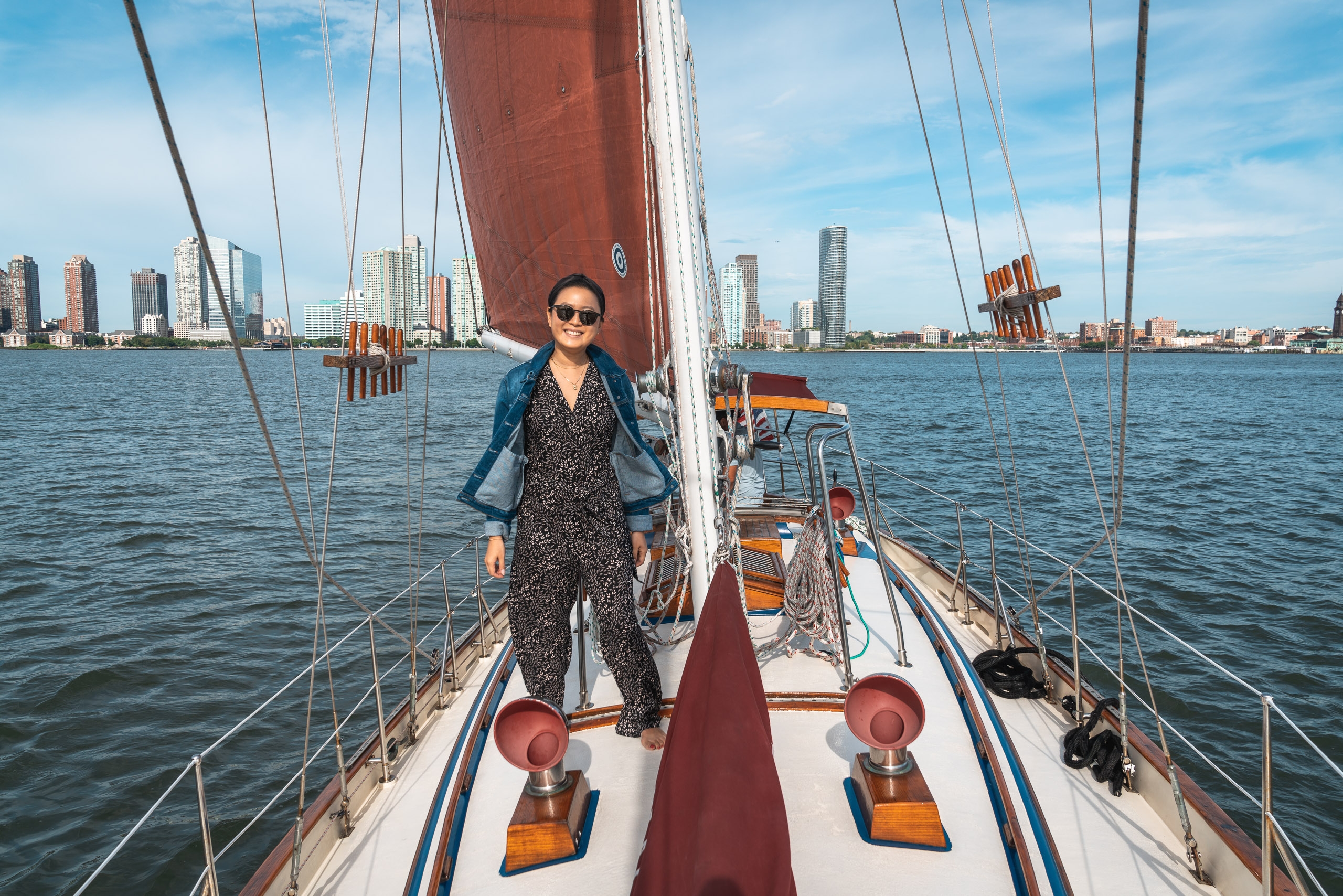 A woman in sunglasses stands on the deck of a sailing vessel with red sails, photographed from above with Manhattan's skyline visible across the blue water behind her