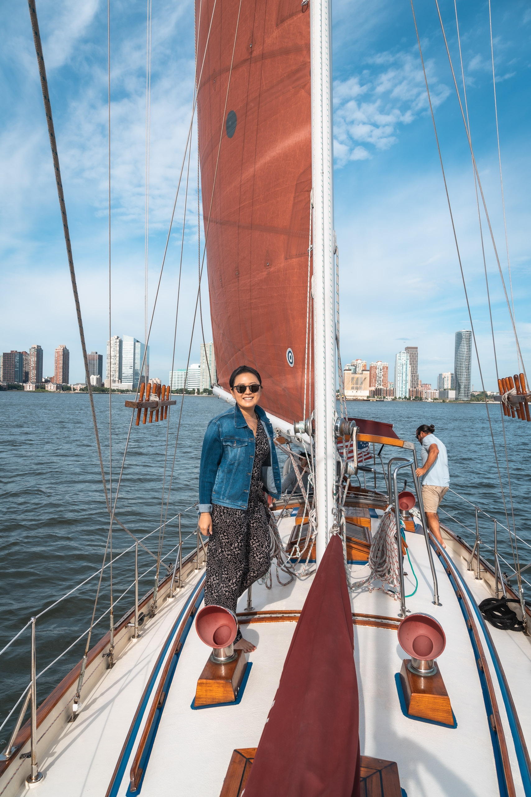 A woman in sunglasses and denim jacket stands on a sailboat's deck with a red sail raised, Manhattan's skyline visible across the blue water behind her.