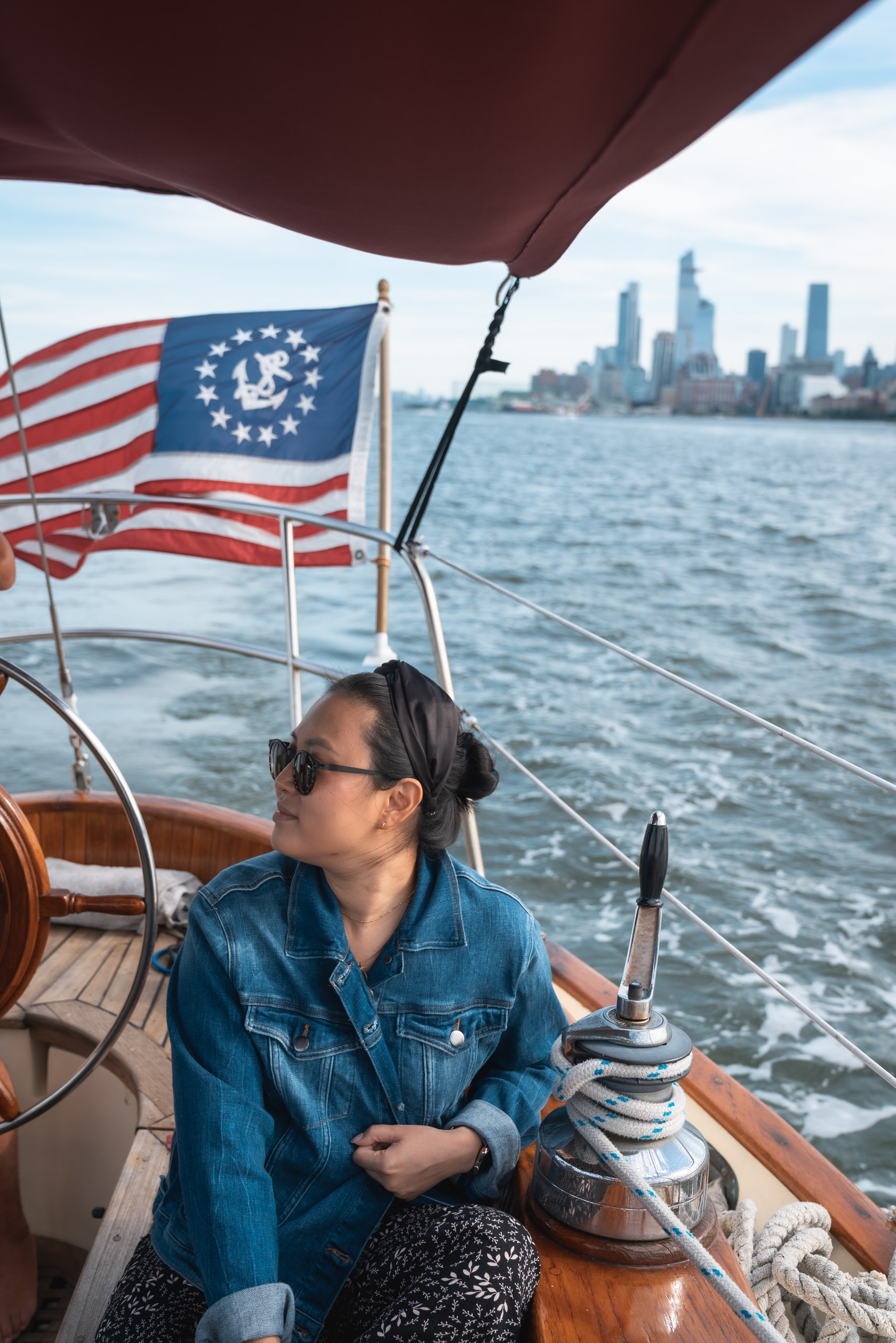 A woman in a denim shirt and sunglasses sits at the helm of a sailboat with the Chicago flag and American flag visible, gazing toward the Manhattan skyline across the water
