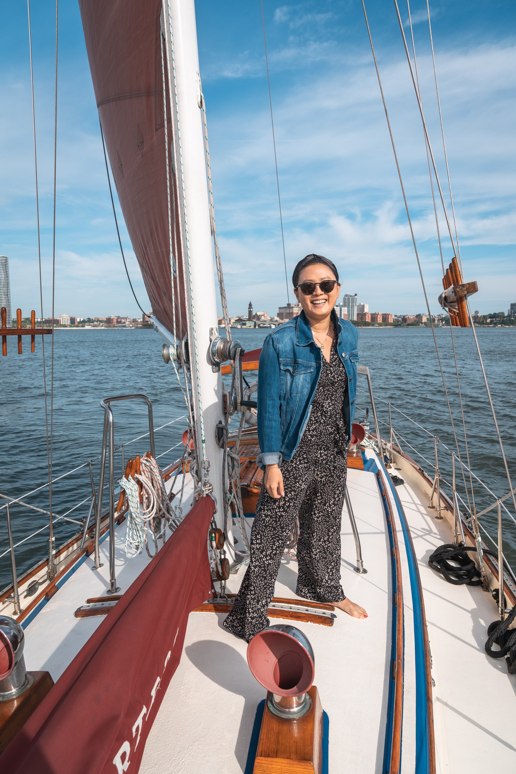 A woman in sunglasses and denim jacket stands on a sailing yacht's deck with billowed sails and Manhattan's skyline visible across the blue water behind her