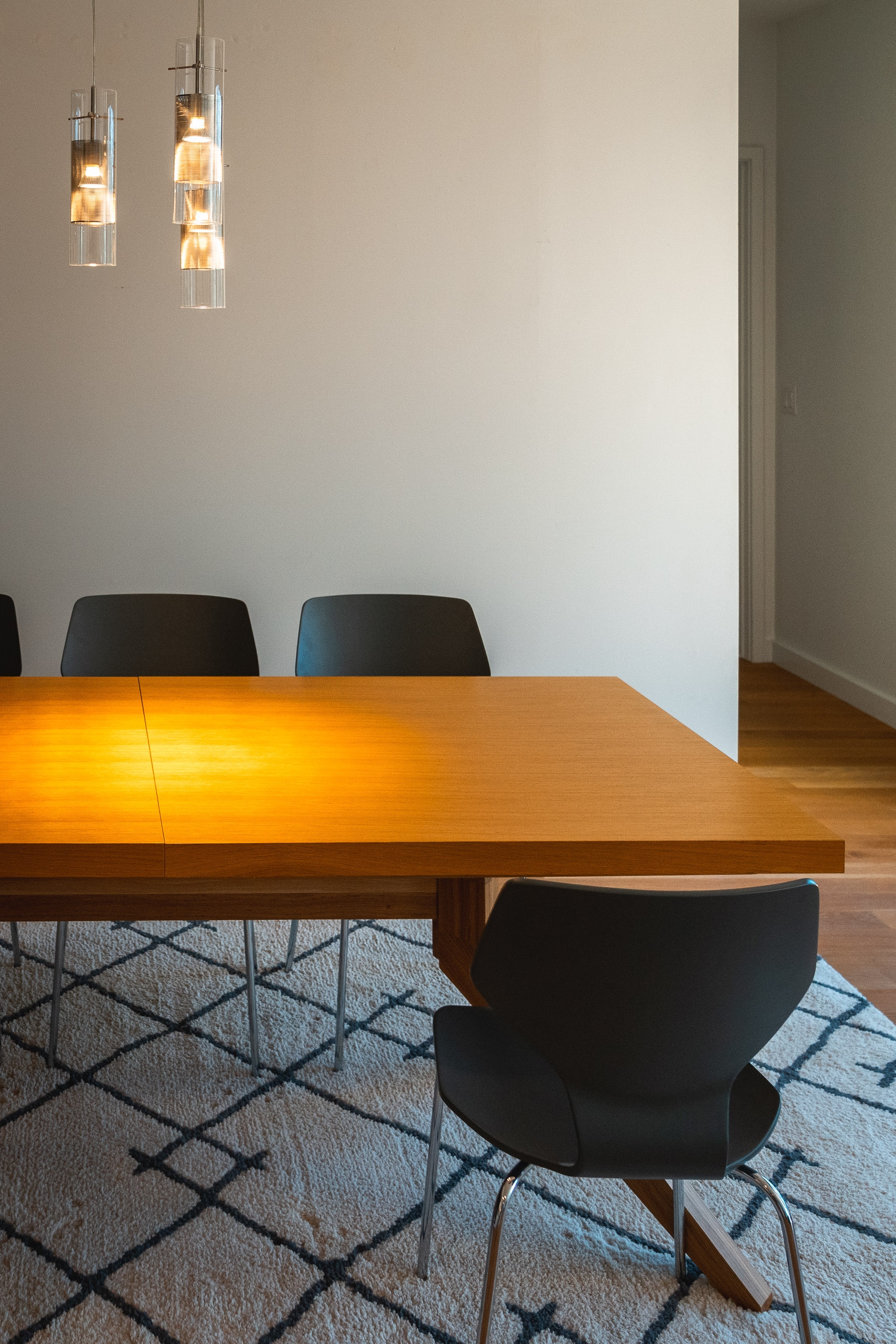 A minimalist dining room features a wooden table with black chairs arranged on a geometric patterned rug, illuminated by pendant lights hanging above in a bright, neutral space