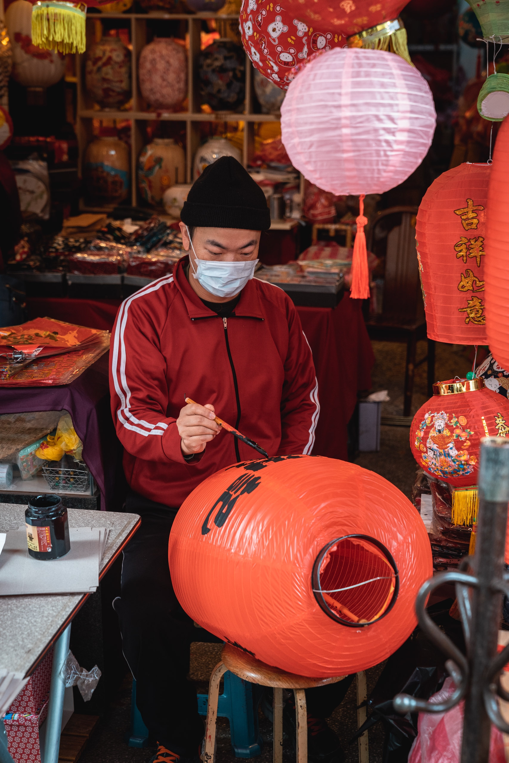A masked artisan in a red jacket carefully paints a large red paper lantern in a shop decorated with traditional Chinese festival ornaments and hanging decorations