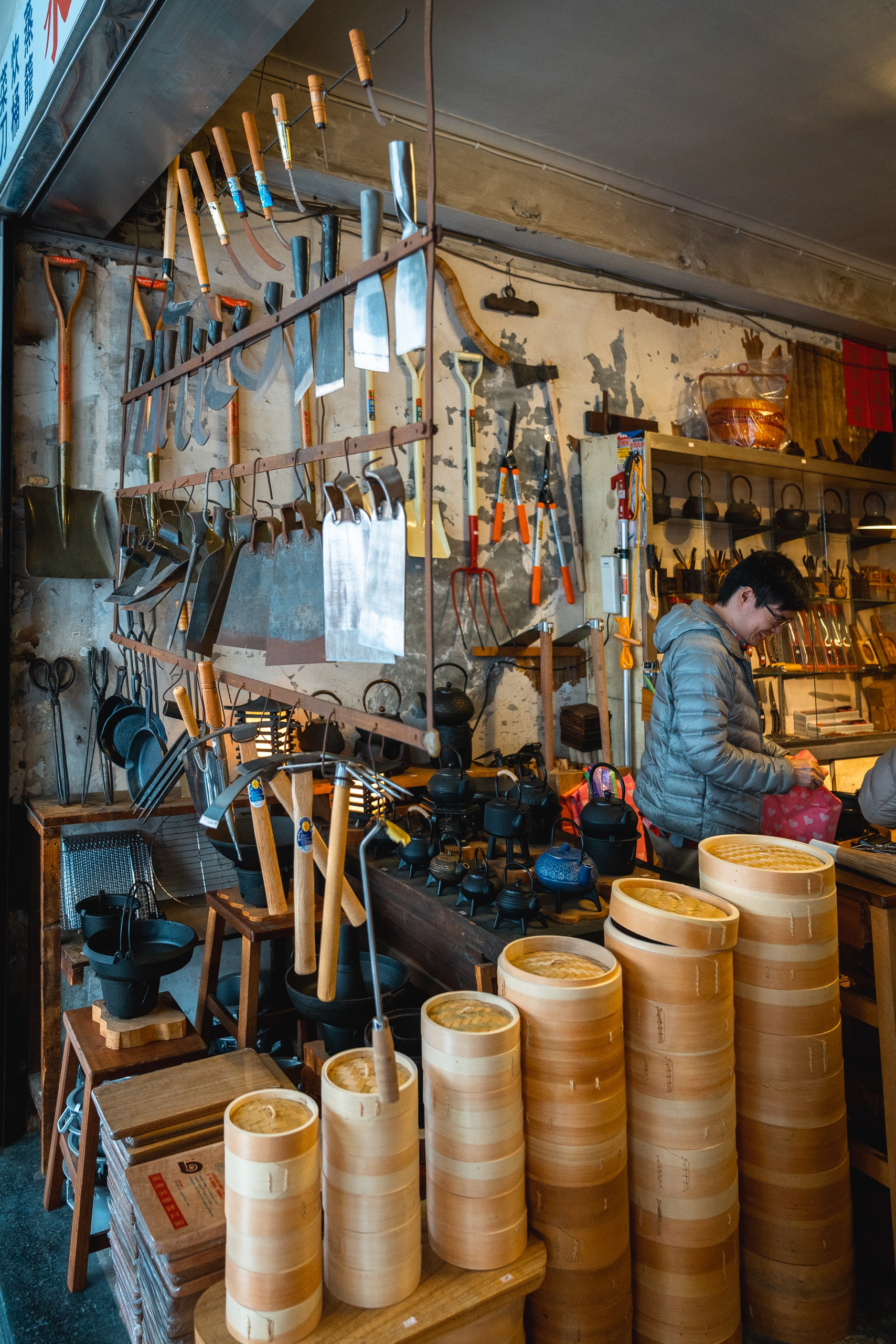 A cluttered artisan workshop displays bamboo steamers in the foreground while hand tools hang organized on weathered walls, with a worker in a blue jacket tending to the shop's interior space