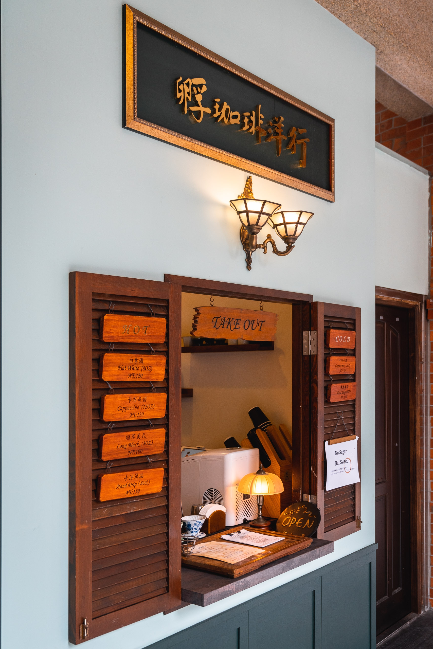 A small wooden takeout window of an Asian restaurant displays Chinese characters on a black signboard above, with warm brass sconces flanking the opening and orange wooden order tickets visible inside the counter