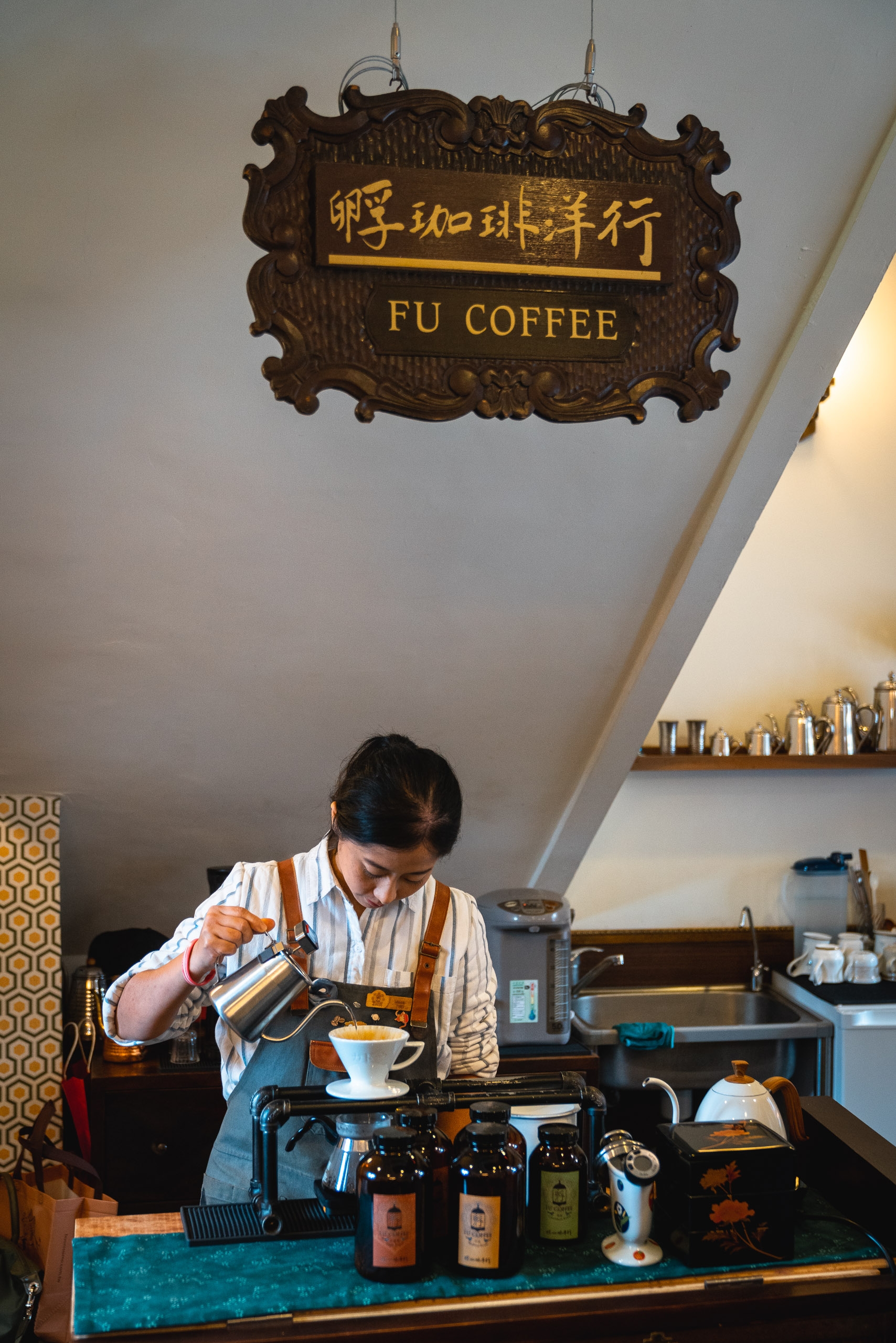 A barista at Fu Coffee shop in Taipei pours hot water from a gooseneck kettle into a filter cone, with Chinese and English signage visible overhead and shelves of supplies behind her