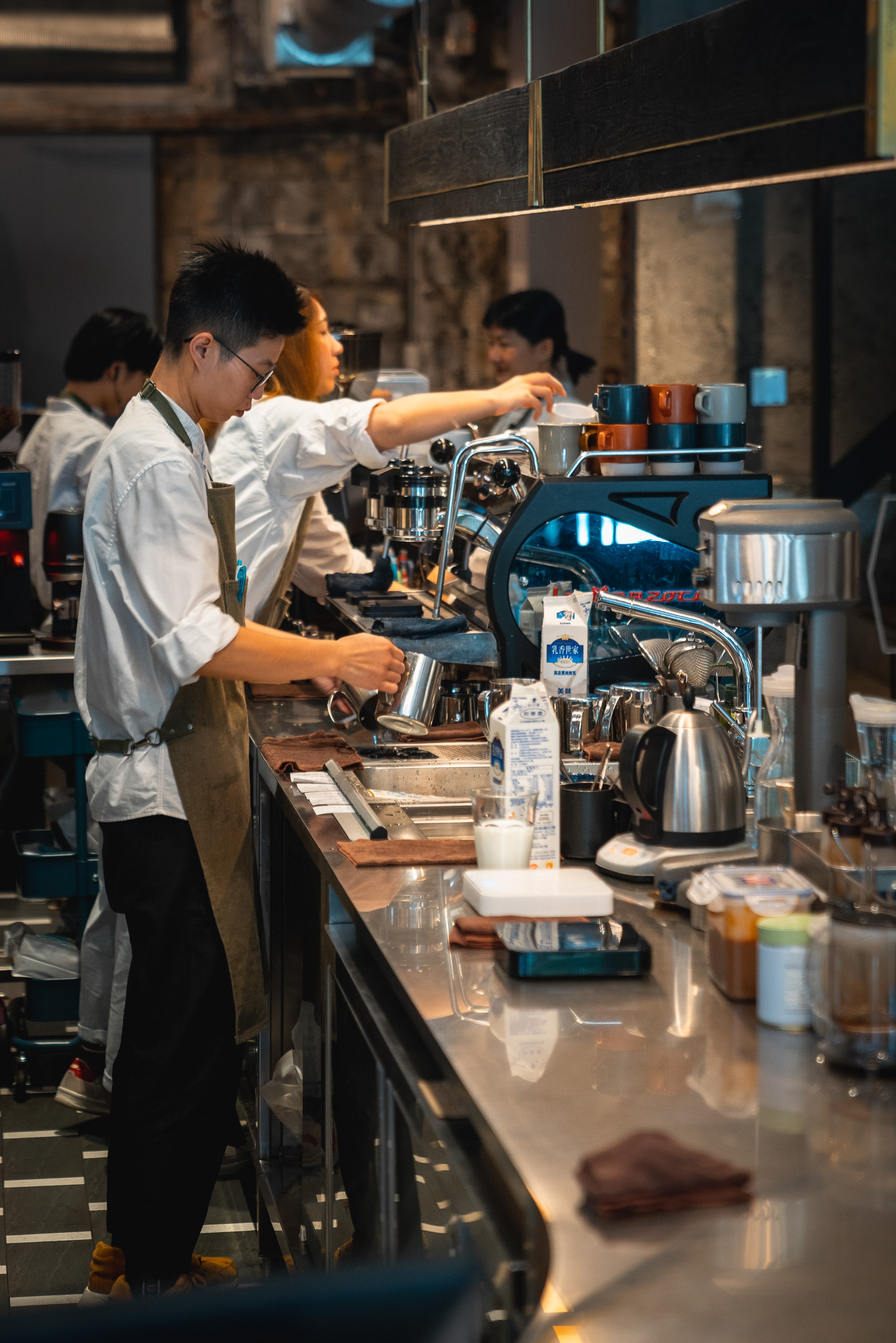 A barista in a white shirt operates an espresso machine at a modern café counter, reaching toward colorful cups while warm lighting illuminates the industrial-style workspace
