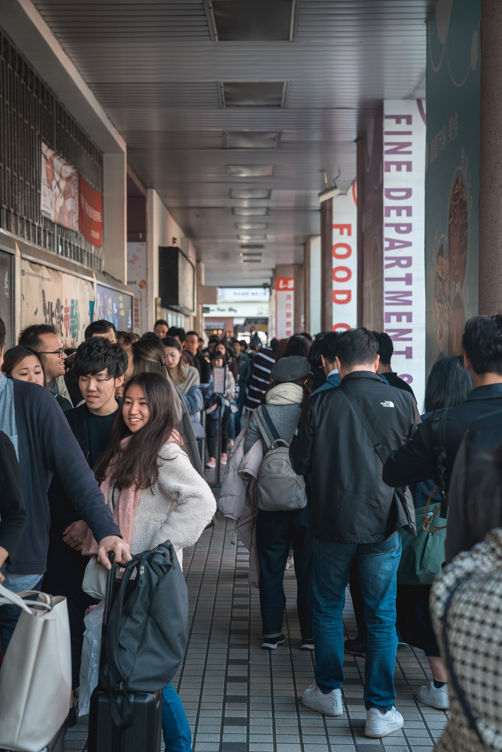 A crowded indoor corridor filled with pedestrians walking past vendor stalls, with red and white signage visible along the metallic ceiling and industrial lighting overhead