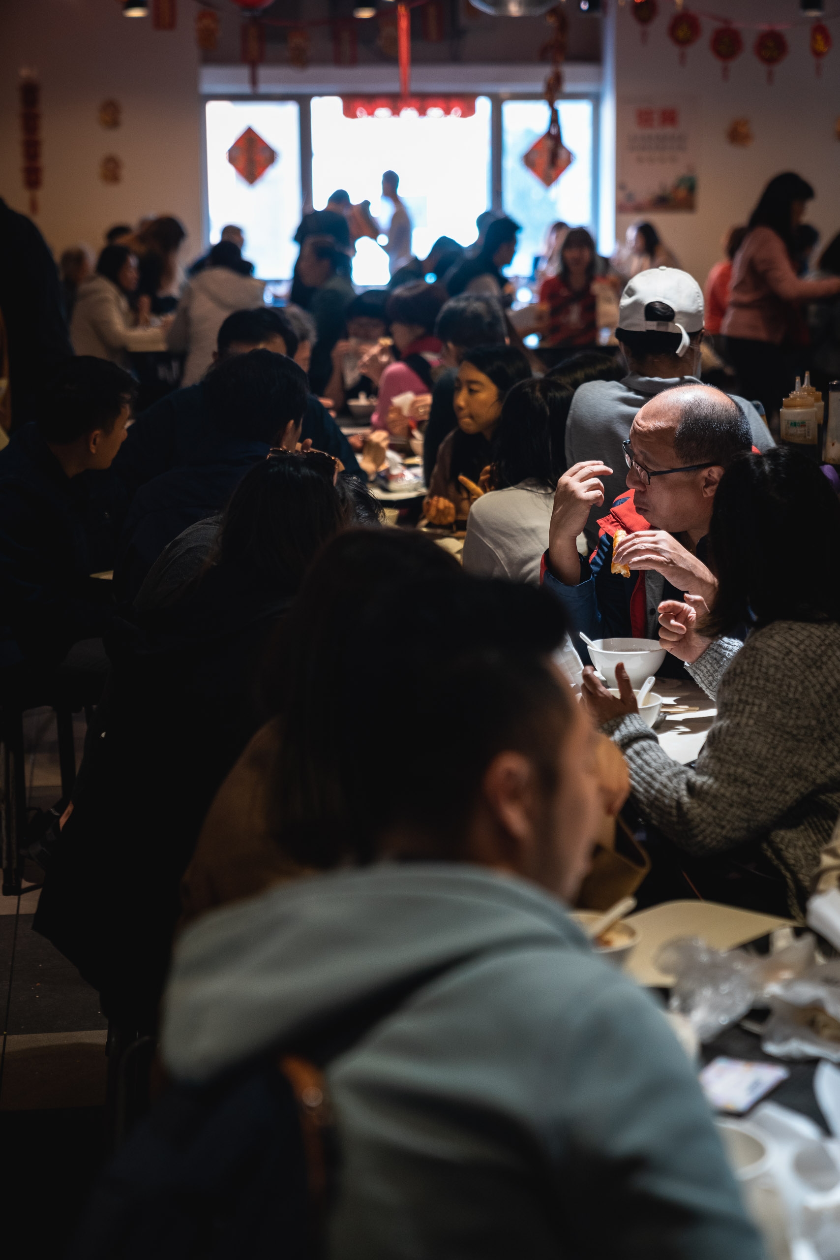 A crowded indoor restaurant or gathering space filled with diners eating at tables, with bright windows and red decorative lanterns visible in the warm, bustling atmosphere