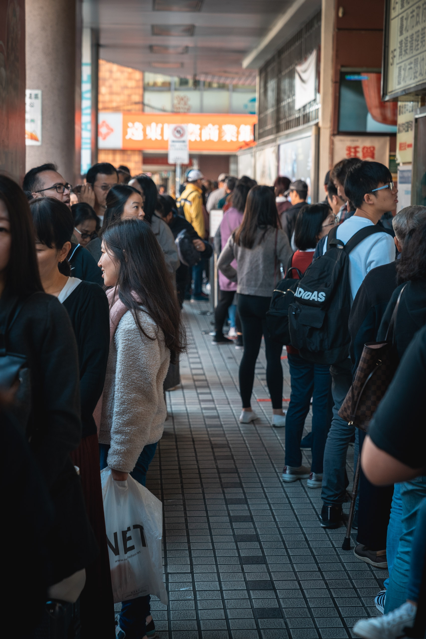 A crowded indoor shopping arcade in Taipei fills with pedestrians walking through a narrow corridor beneath bright orange signage and fluorescent lighting