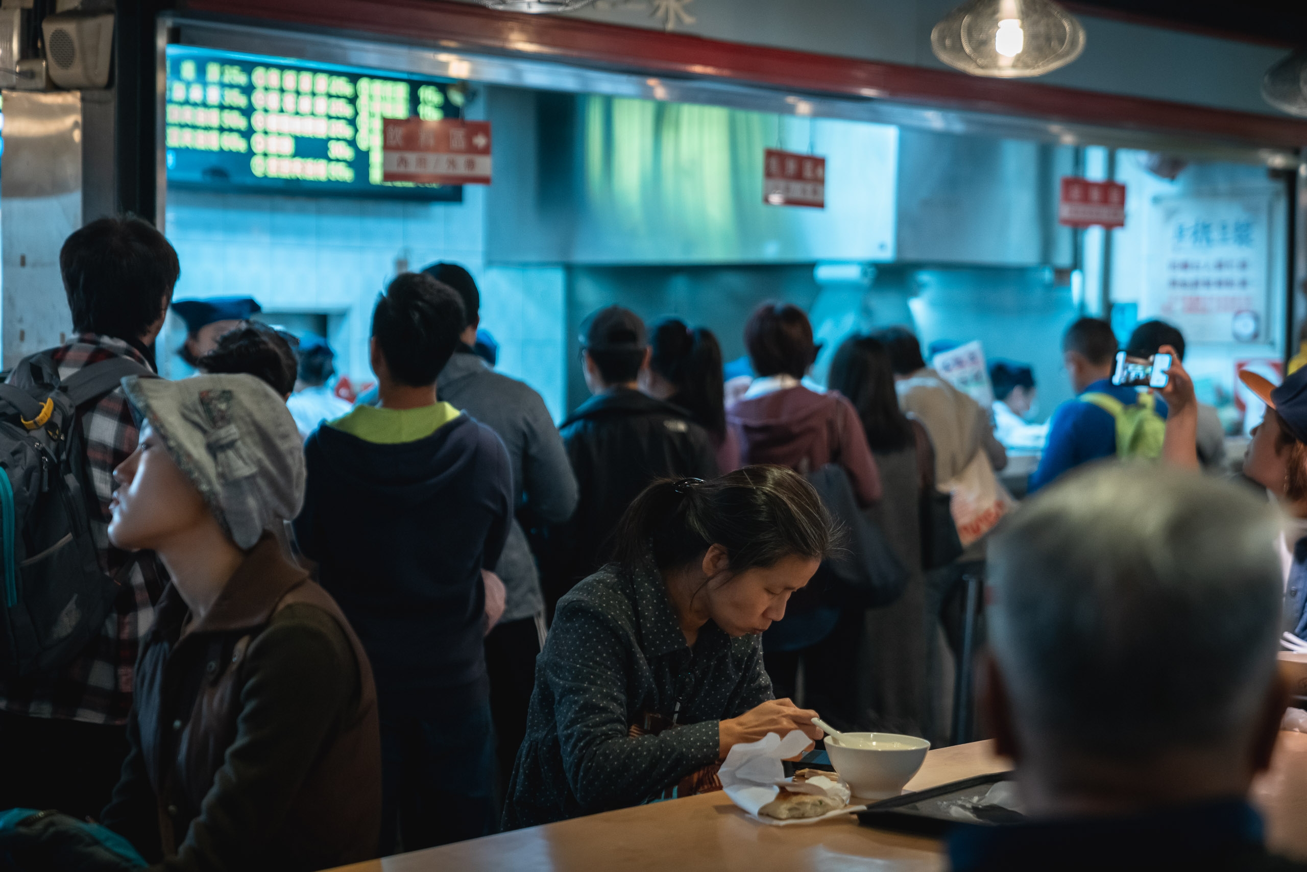 A crowded Asian food counter with customers waiting in line while a woman in the foreground eats noodles at a table, illuminated by bright blue overhead lighting and menu boards