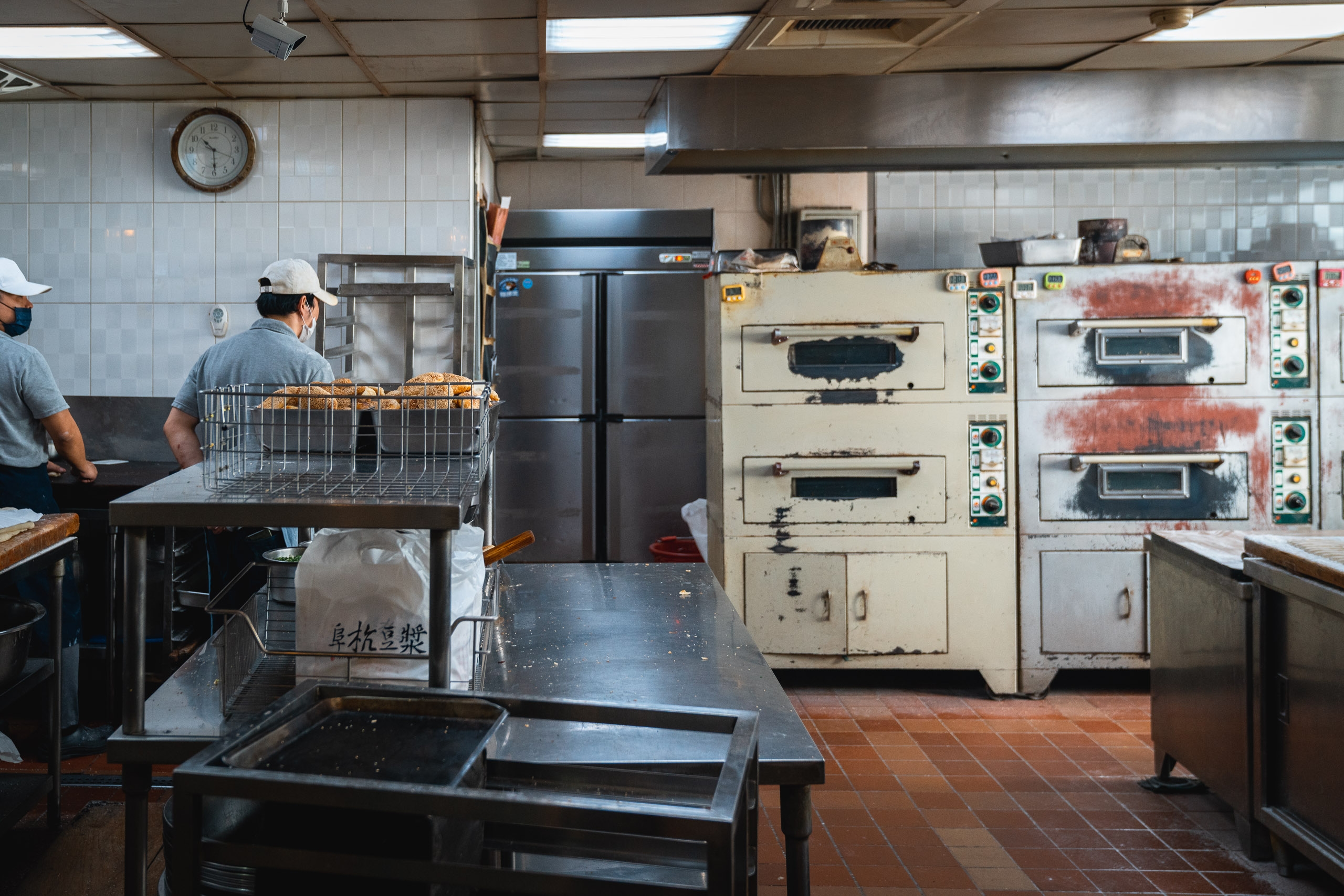 A commercial kitchen in Taipei features industrial ovens and cooking equipment with workers in caps preparing food, fluorescent ceiling lights casting even illumination across the tiled walls and terracotta floor