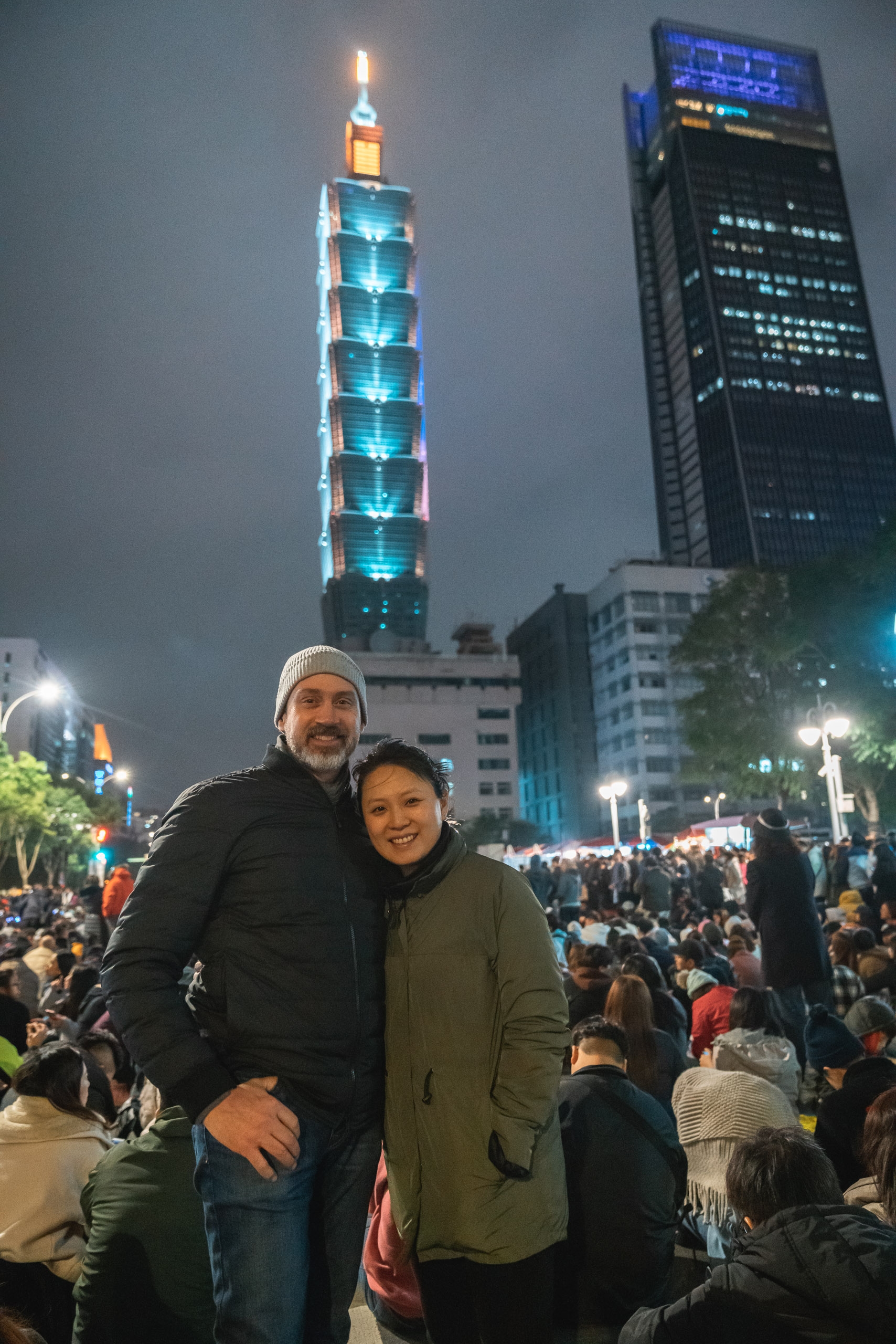 A couple poses together in a crowded nighttime plaza beneath illuminated skyscrapers, with Taipei 101's glowing turquoise tower rising prominently in the background