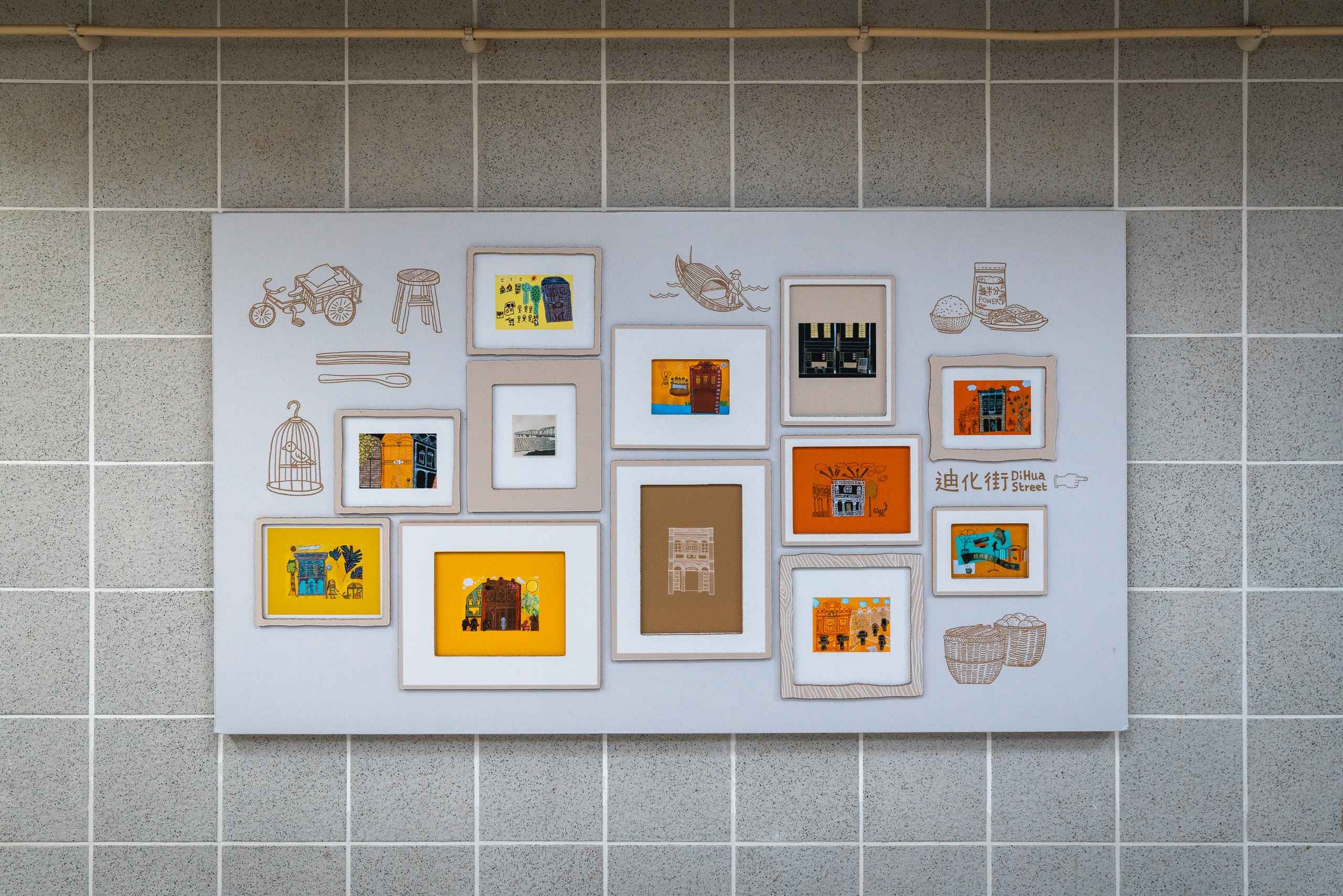 A white gallery wall display mounted on gray tile features an eclectic arrangement of framed prints, drawings, and photographs in warm earth tones and vibrant orange and yellow hues, depicting architectural and domestic scenes