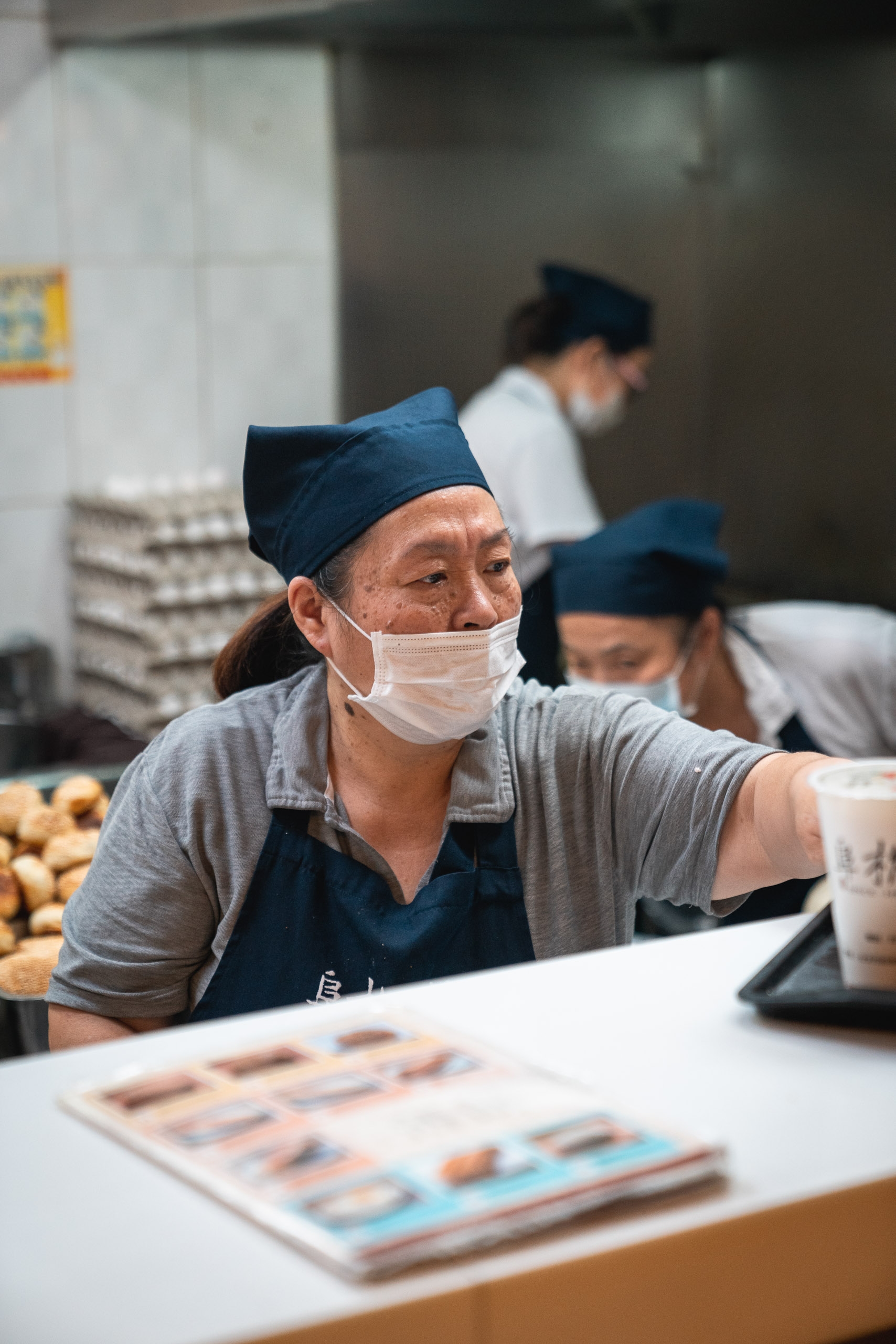 A masked worker in a blue headband and apron stands at a counter in a food preparation facility, with colleagues visible in the blurred background and product images on the workspace surface