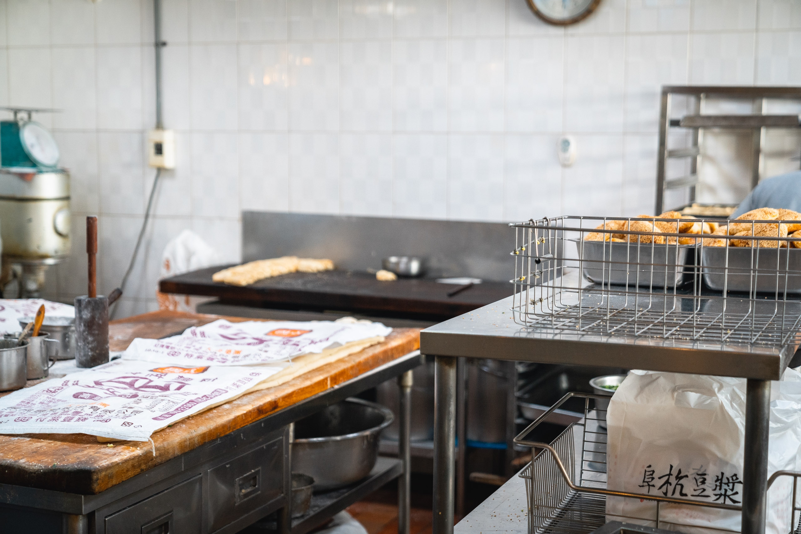 A bakery kitchen workspace displays fresh loaves of bread in wire baskets on stainless steel tables, with baked goods and price labels visible under fluorescent overhead lighting