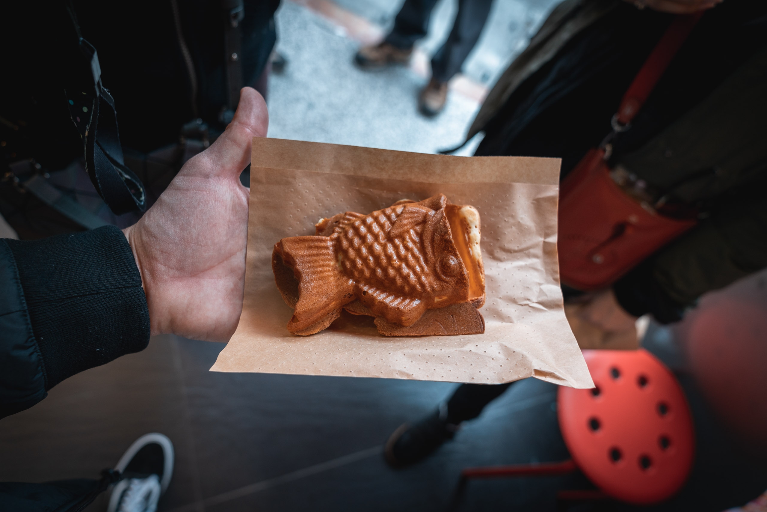 A hand holds a paper wrapper containing a golden-brown fish-shaped pastry, surrounded by people's legs and feet on a grey pavement in daylight