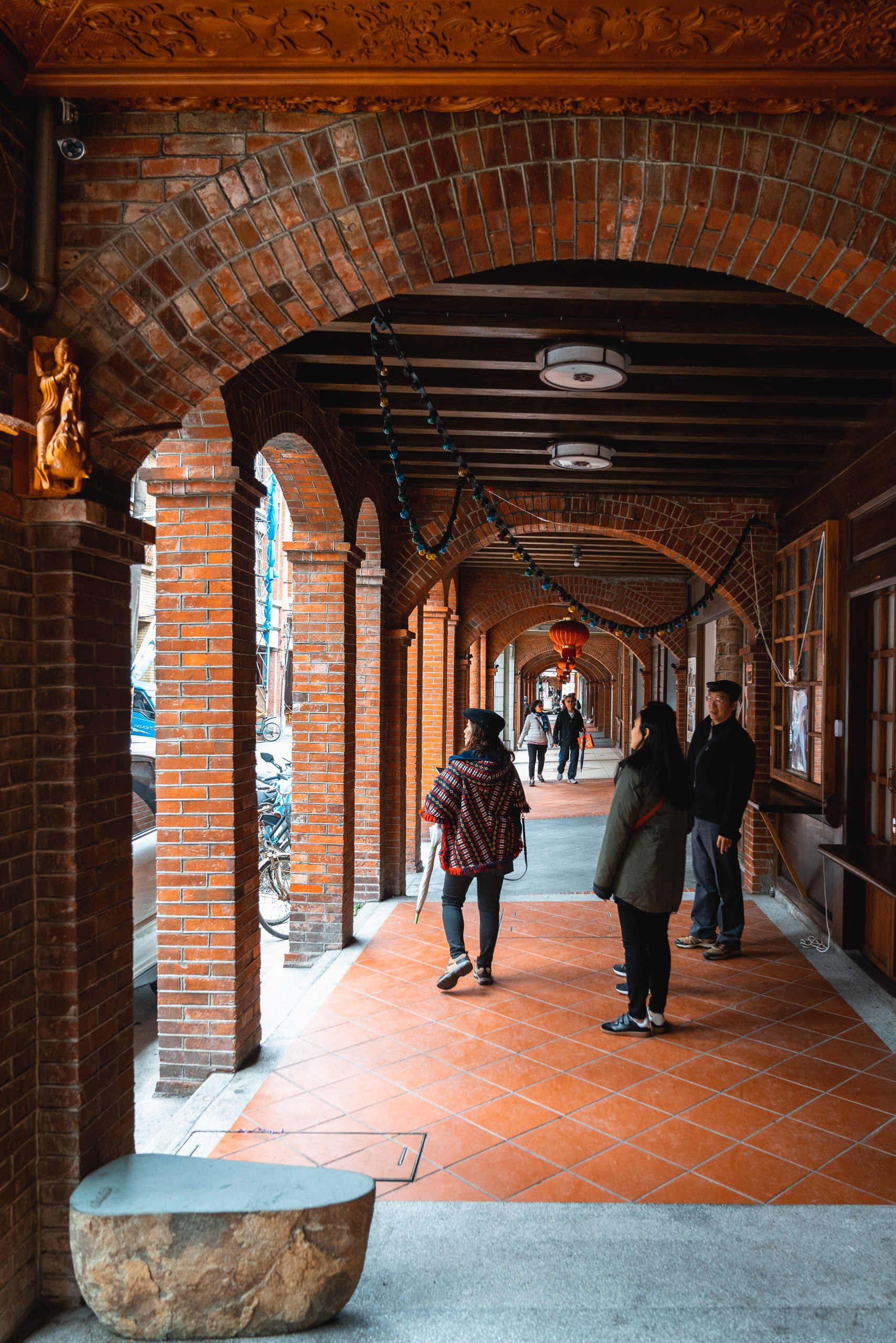 Pedestrians walk through a covered brick archway with decorative wooden beams, warm sunlight streaming through the colonnade in this historic Taipei passage