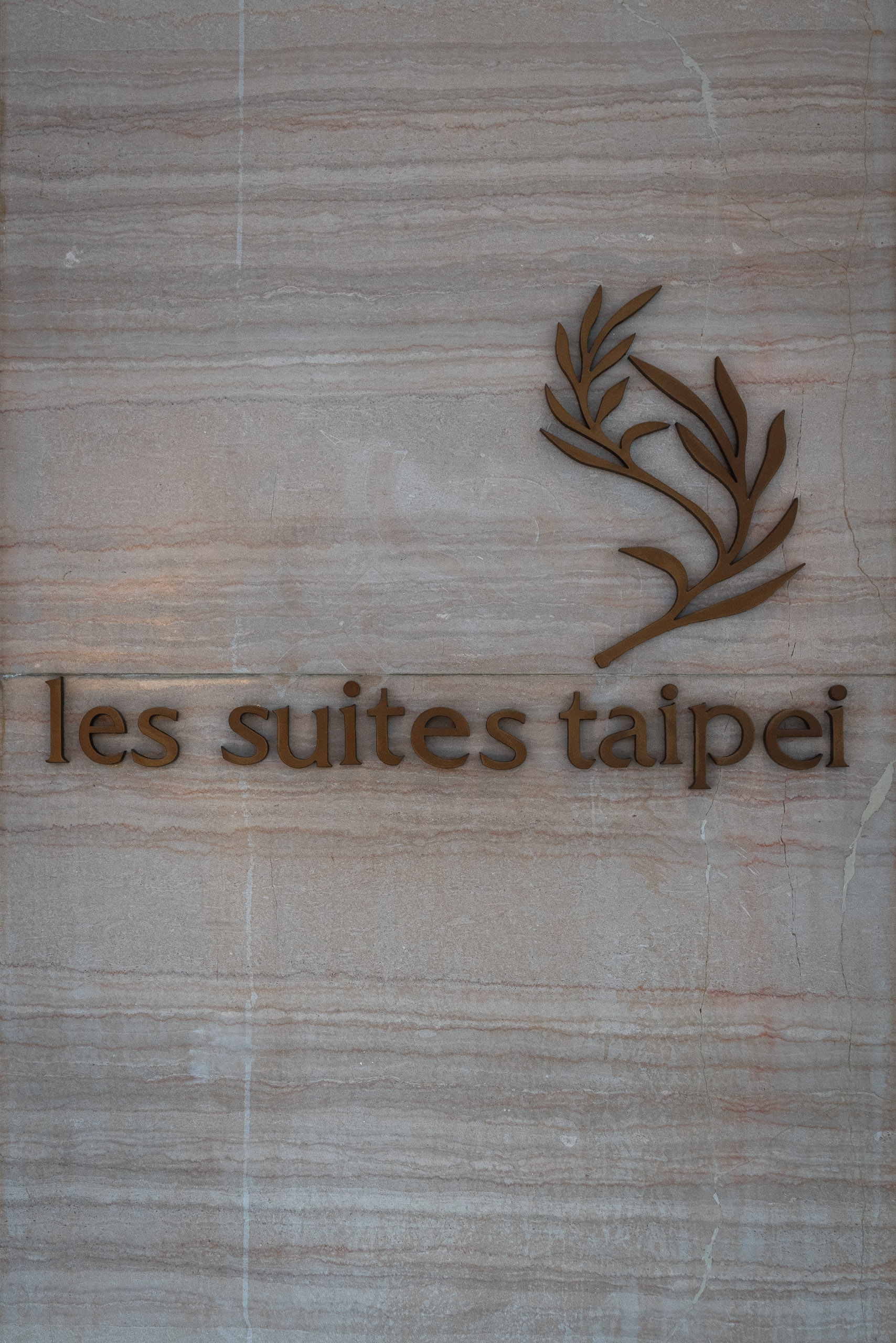 Wooden signage carved with "les suites taipei" text and a stylized branch motif mounted on a light gray textured wall surface