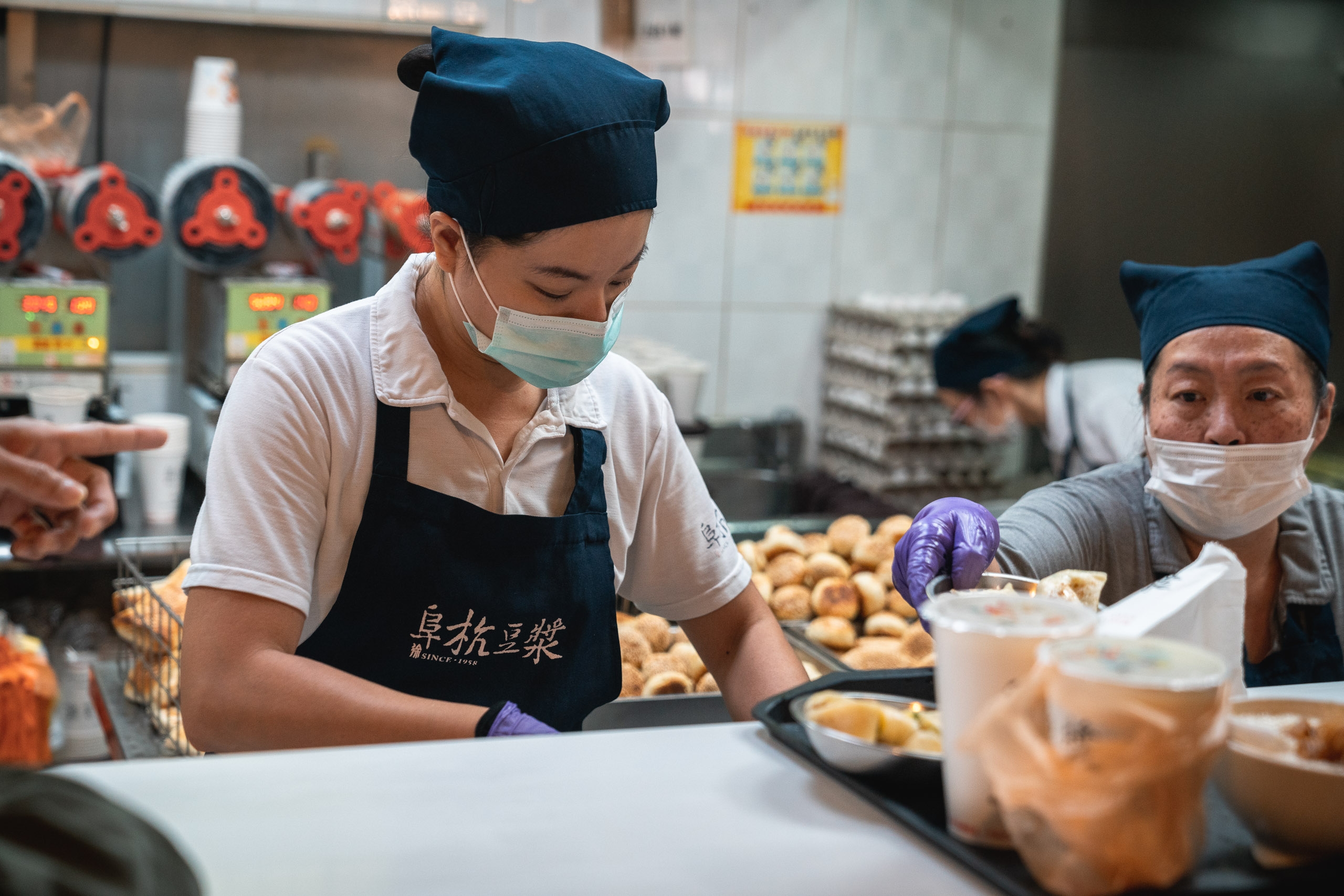 A masked food worker in a blue cap and apron prepares dishes at a busy kitchen counter, with colleagues and golden-brown ingredients visible in the warm-lit background