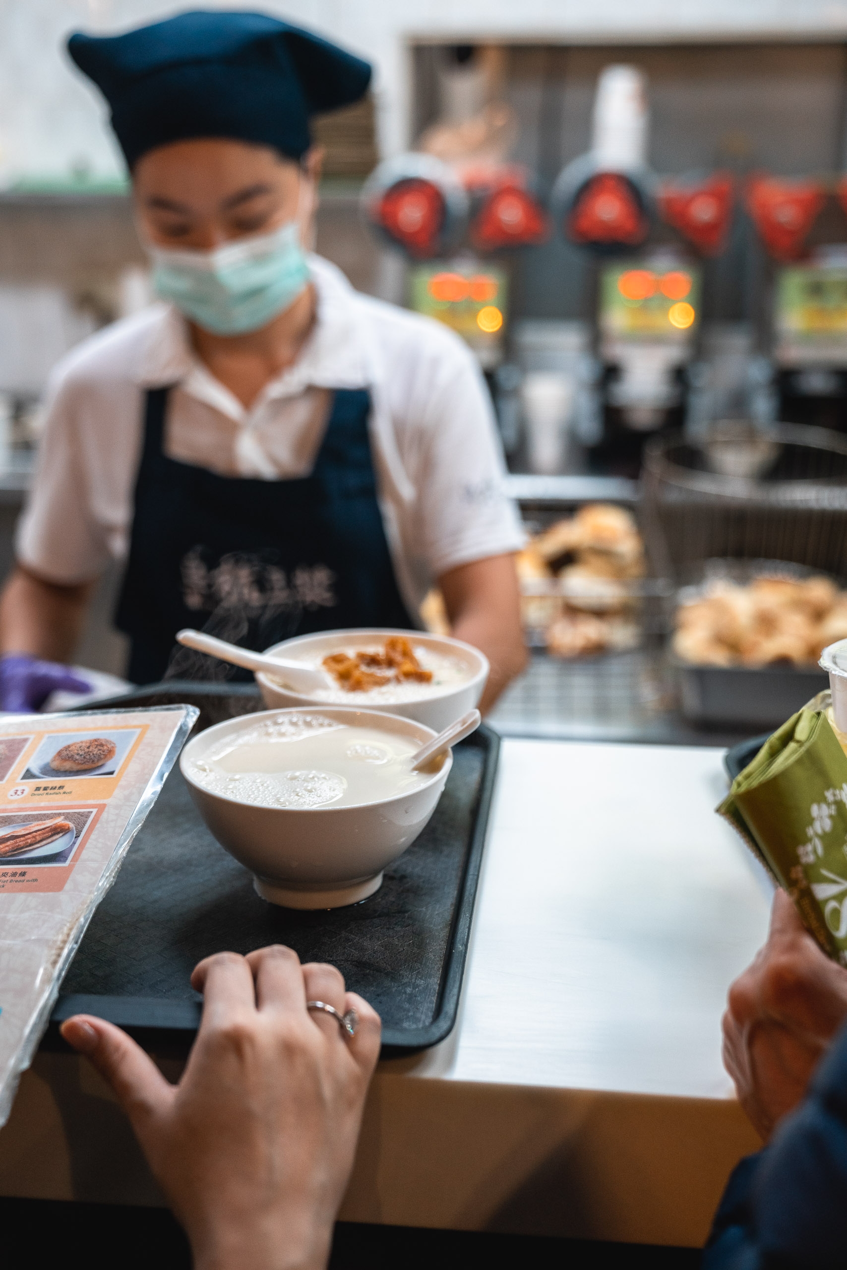 A masked server in a black apron presents a bowl of creamy soup to a customer across a counter in a busy Taipei food establishment, with kitchen equipment visible in the blurred background.
