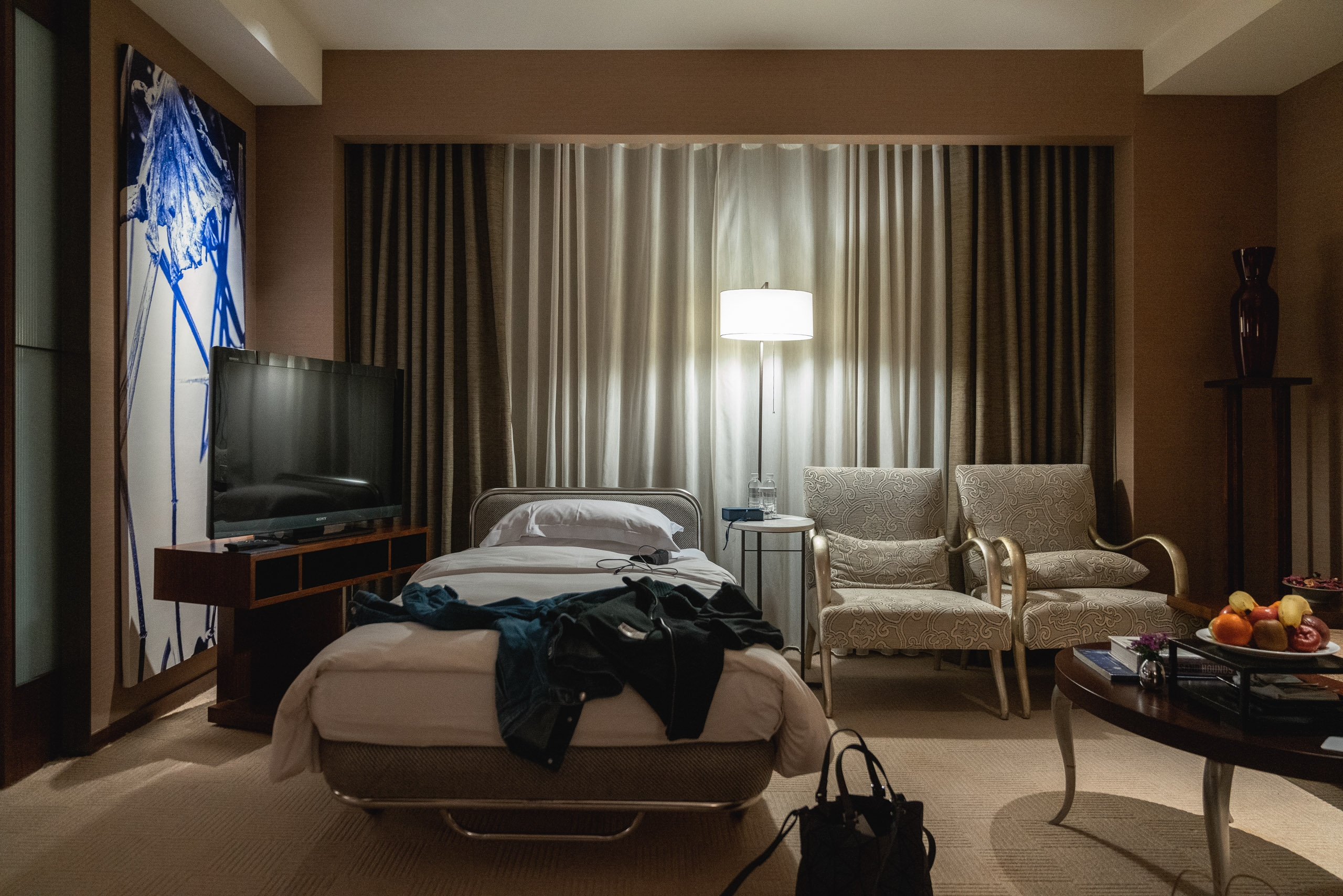 A modern hotel bedroom in warm brown tones features a bed with scattered clothing, contemporary furnishings, floor lamp, and sheer curtains filtering soft light from large windows