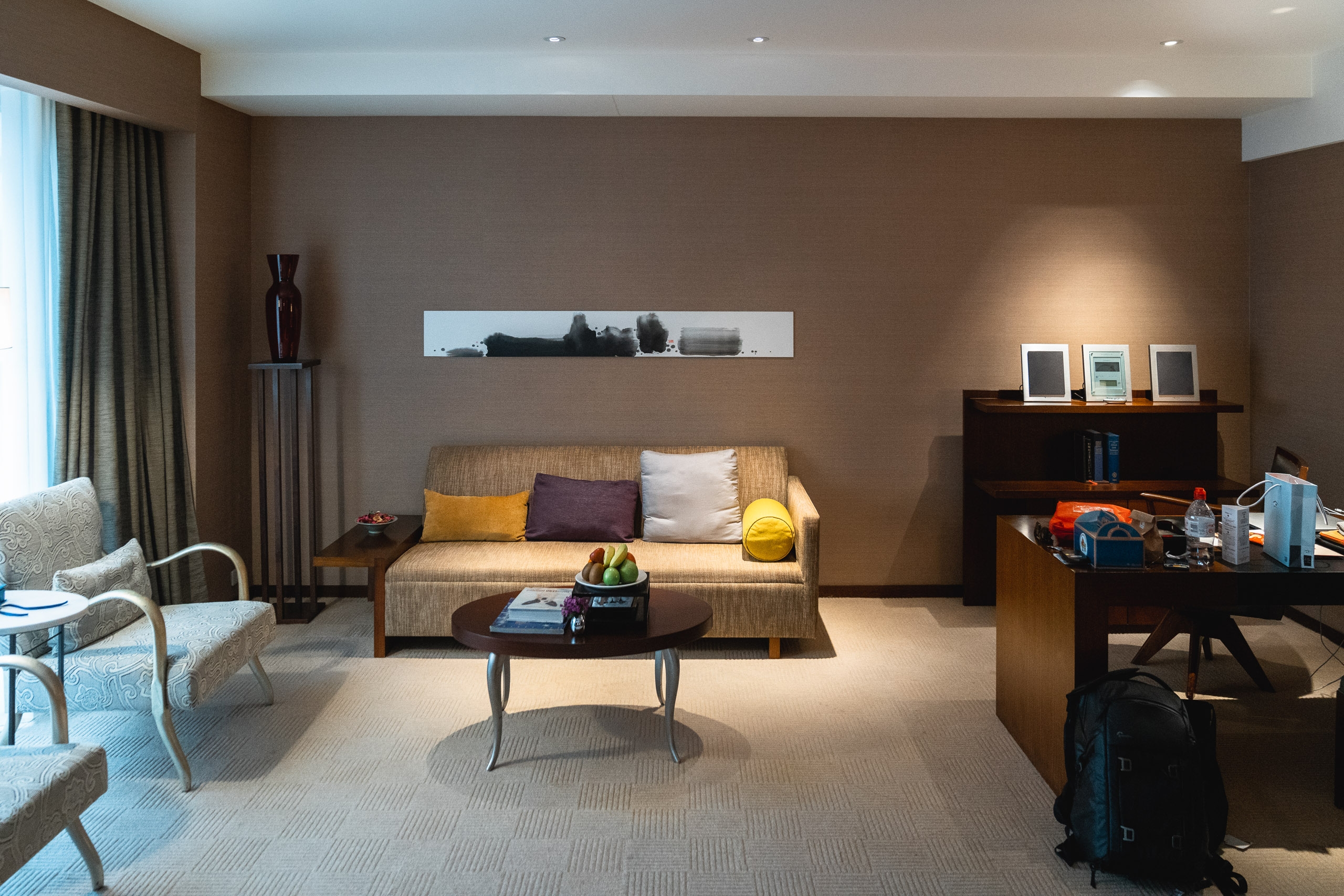 A warm-toned hotel suite features a tan upholstered sofa with colorful pillows facing a dark coffee table, with brown walls, recessed lighting, and modern furniture arranged in a spacious contemporary living area