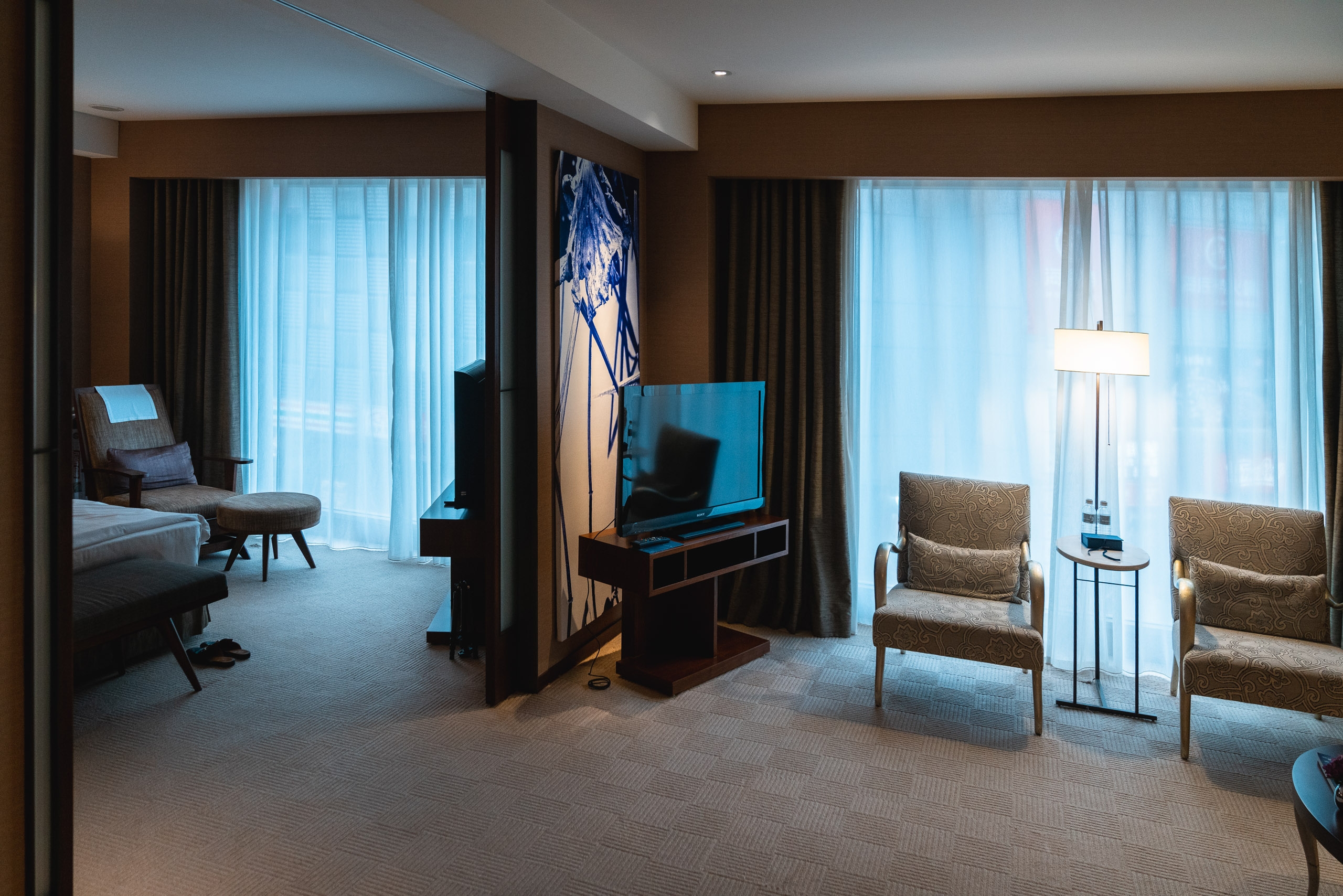 Modern hotel suite with brown walls, two upholstered chairs, television on dark stand, and bright blue-tinted natural light streaming through floor-to-ceiling curtained windows