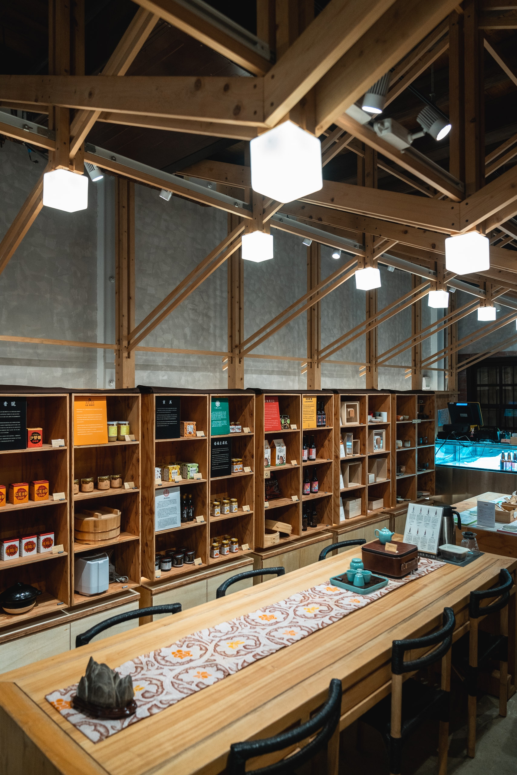 A modern retail space with exposed wooden beam ceiling and pendant lighting displays shelves of packaged products along concrete walls, with a wooden counter in the foreground