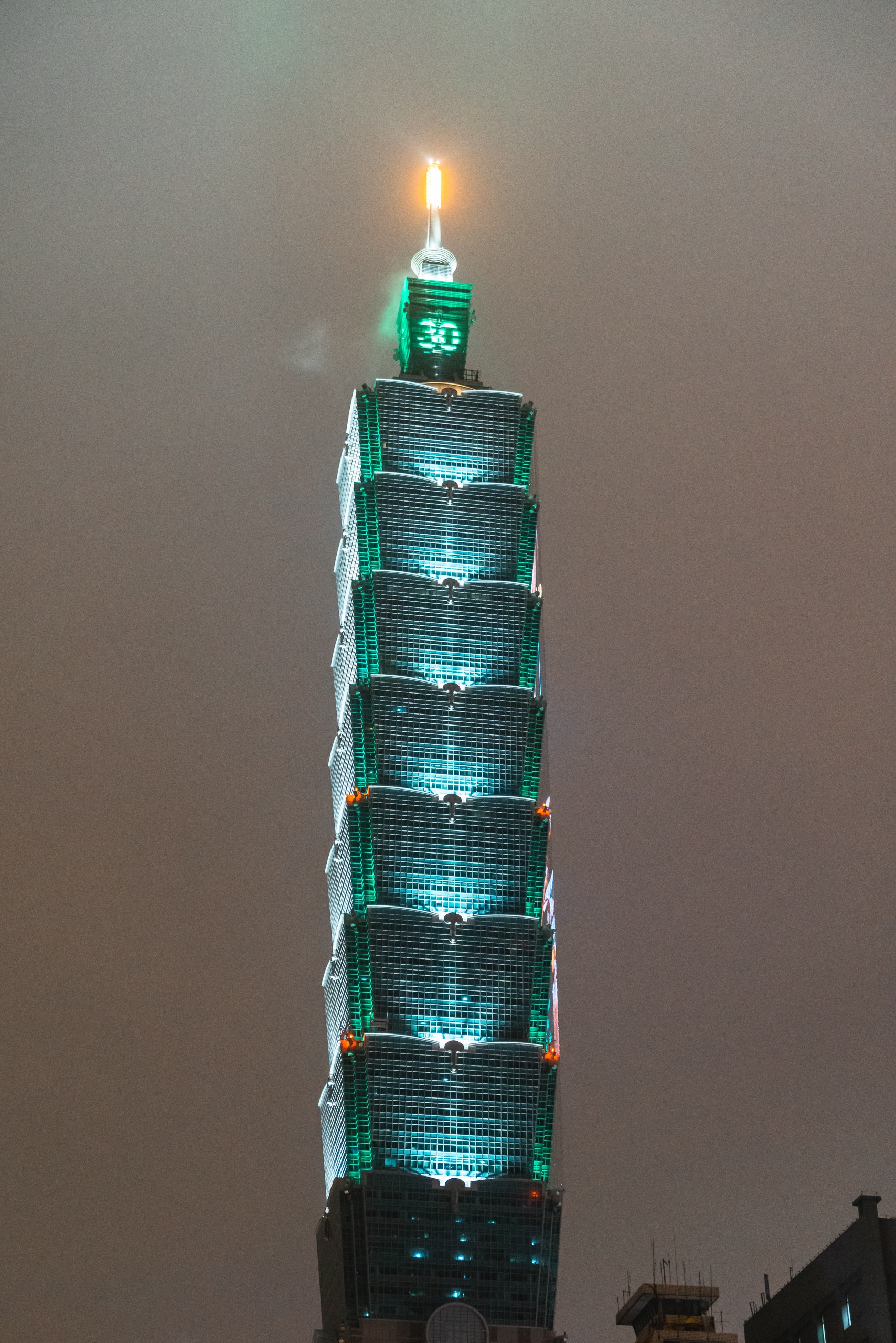 A tall modern skyscraper illuminated with teal and green lights at night, its distinctive segmented sections stacked vertically, crowned with a glowing golden spire piercing the hazy sky above the city