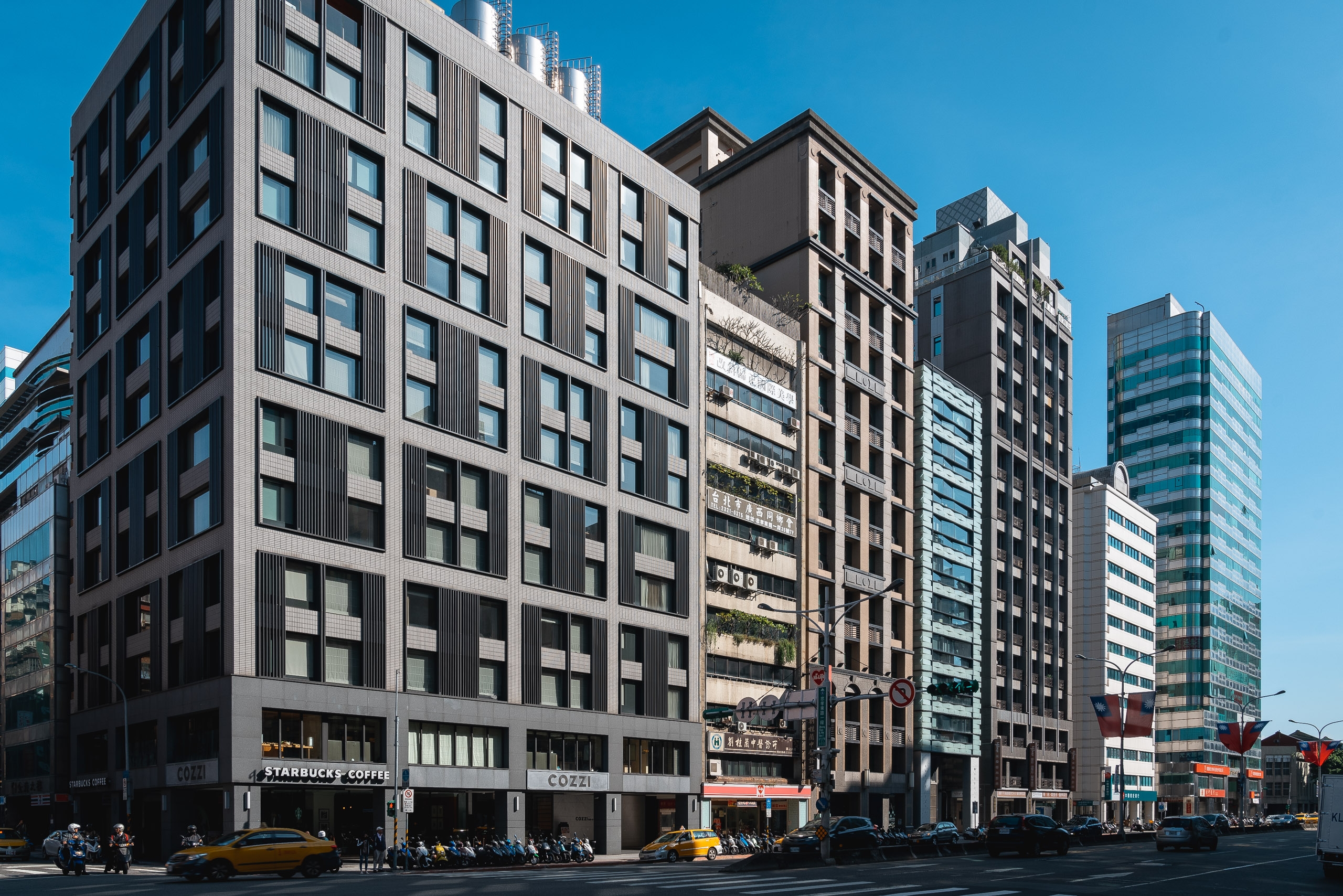 A row of modern mixed-use buildings with black and gray facades lines a busy urban street under clear blue sky, with ground-floor retail including a visible Starbucks and yellow taxis passing by