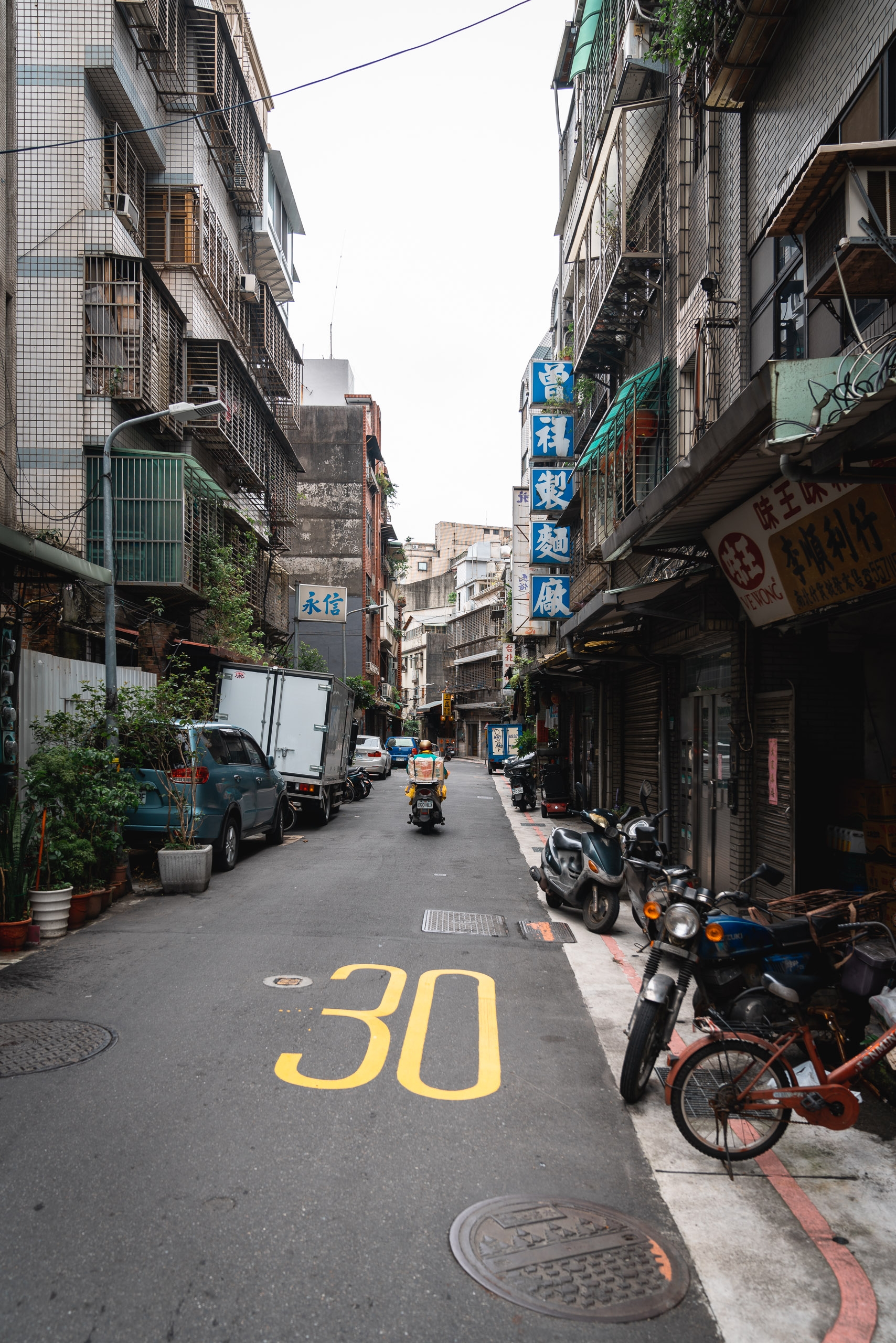A narrow urban alley in Taipei lined with weathered buildings, parked motorcycles, and Chinese signage, with yellow "30" road markings visible on the pavement under overcast sky