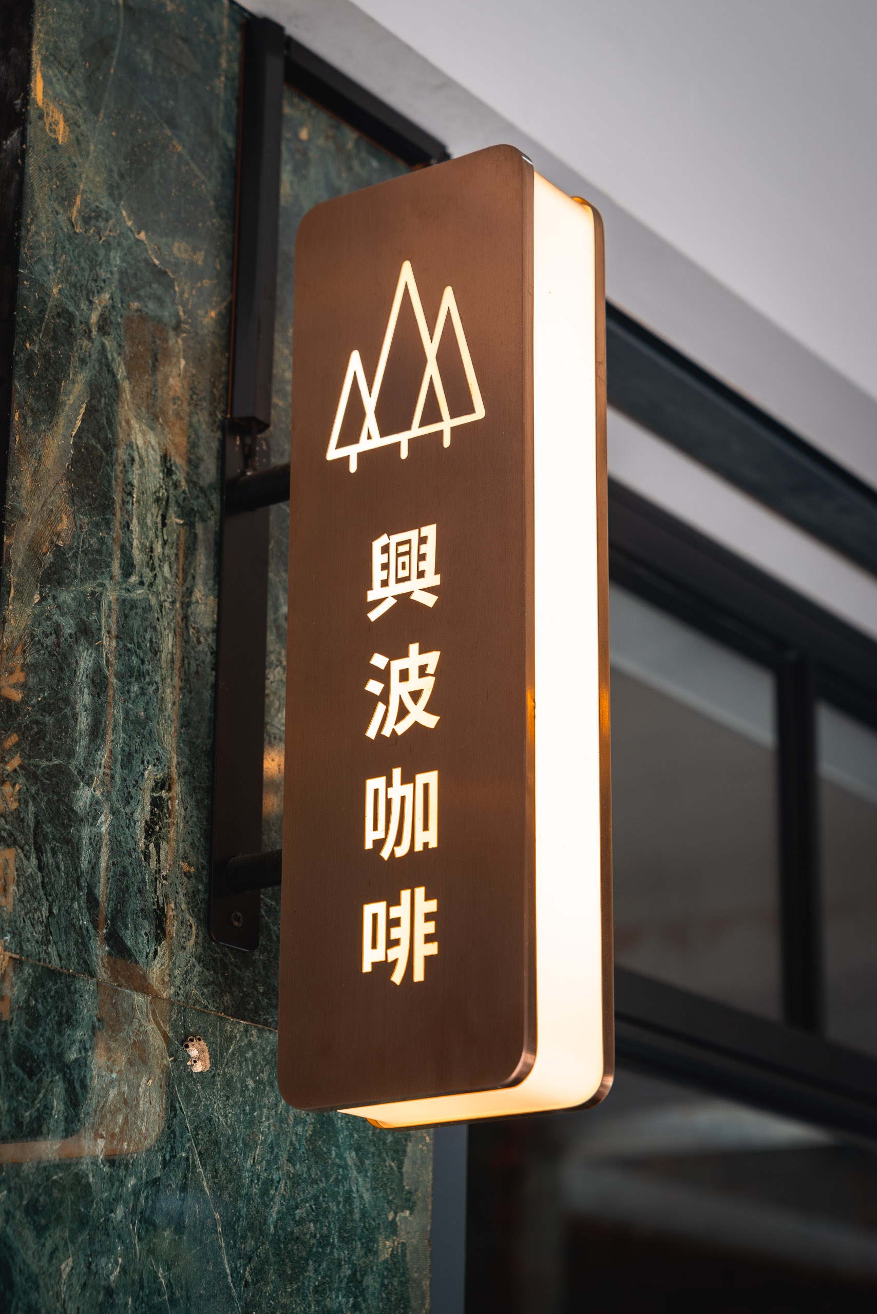 A vertical illuminated brown signage with white neon tree icon and Chinese characters mounted on a weathered teal-green stone wall beside a storefront window