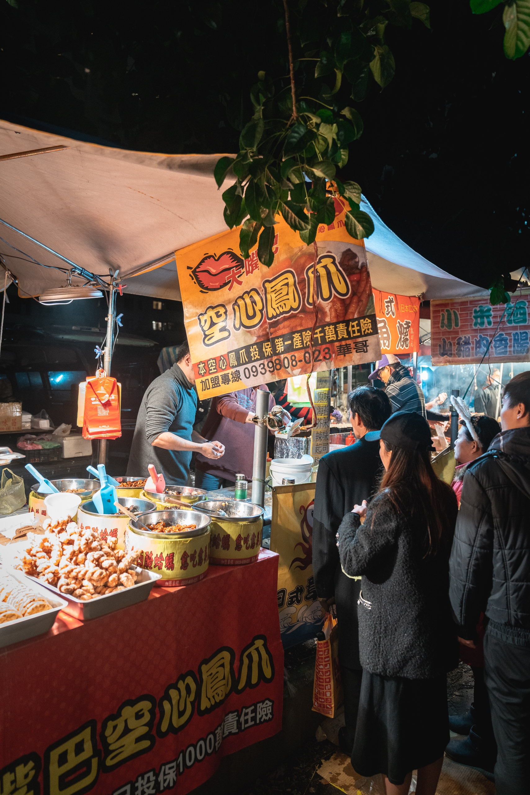 A night market food vendor stall with orange signage and red banner displays grilled skewers to customers gathered under warm tent lighting, with climbing vines overhead and adjacent shops visible