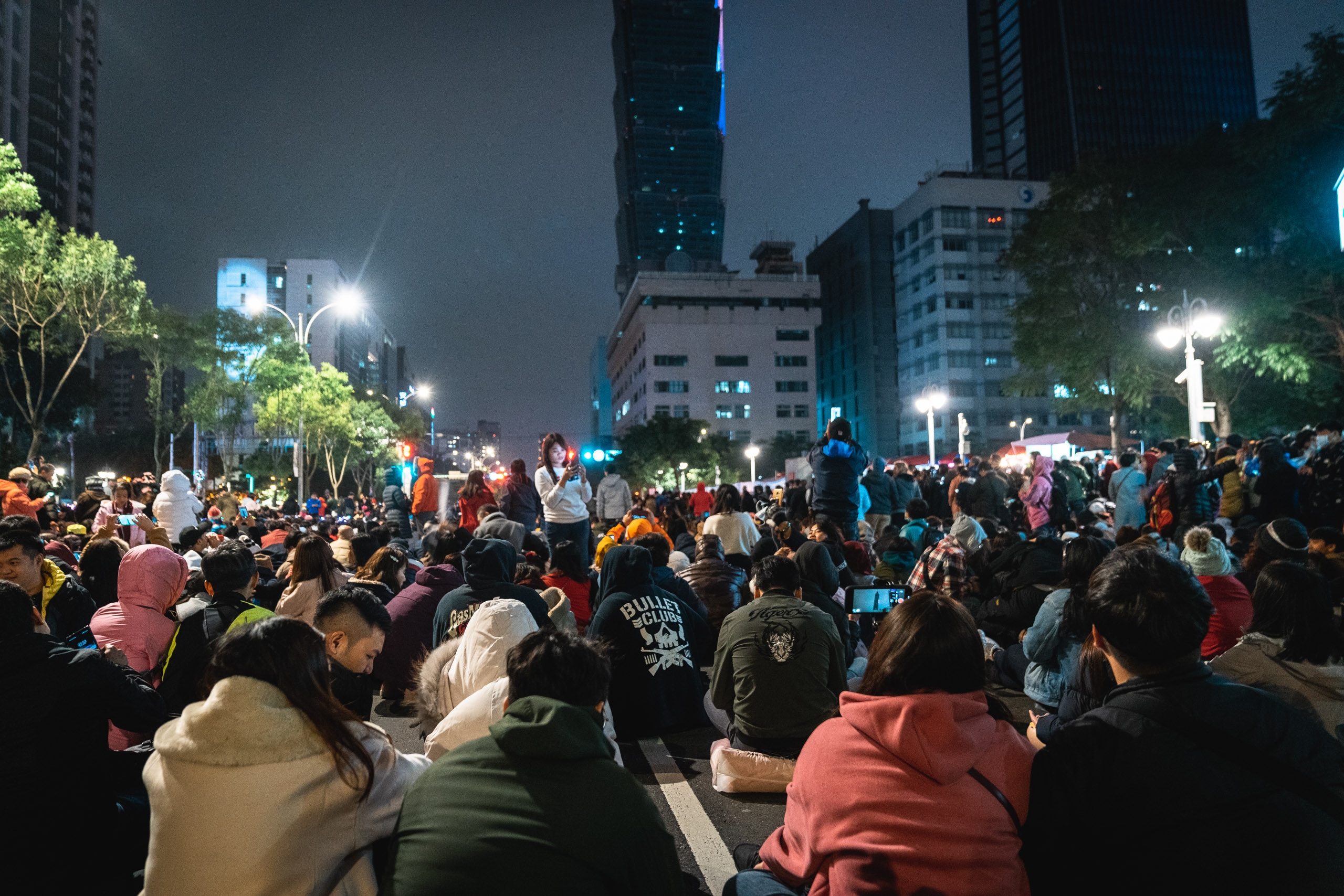 A large nighttime crowd gathers in an urban plaza surrounded by illuminated high-rise buildings and glowing street trees in Taipei