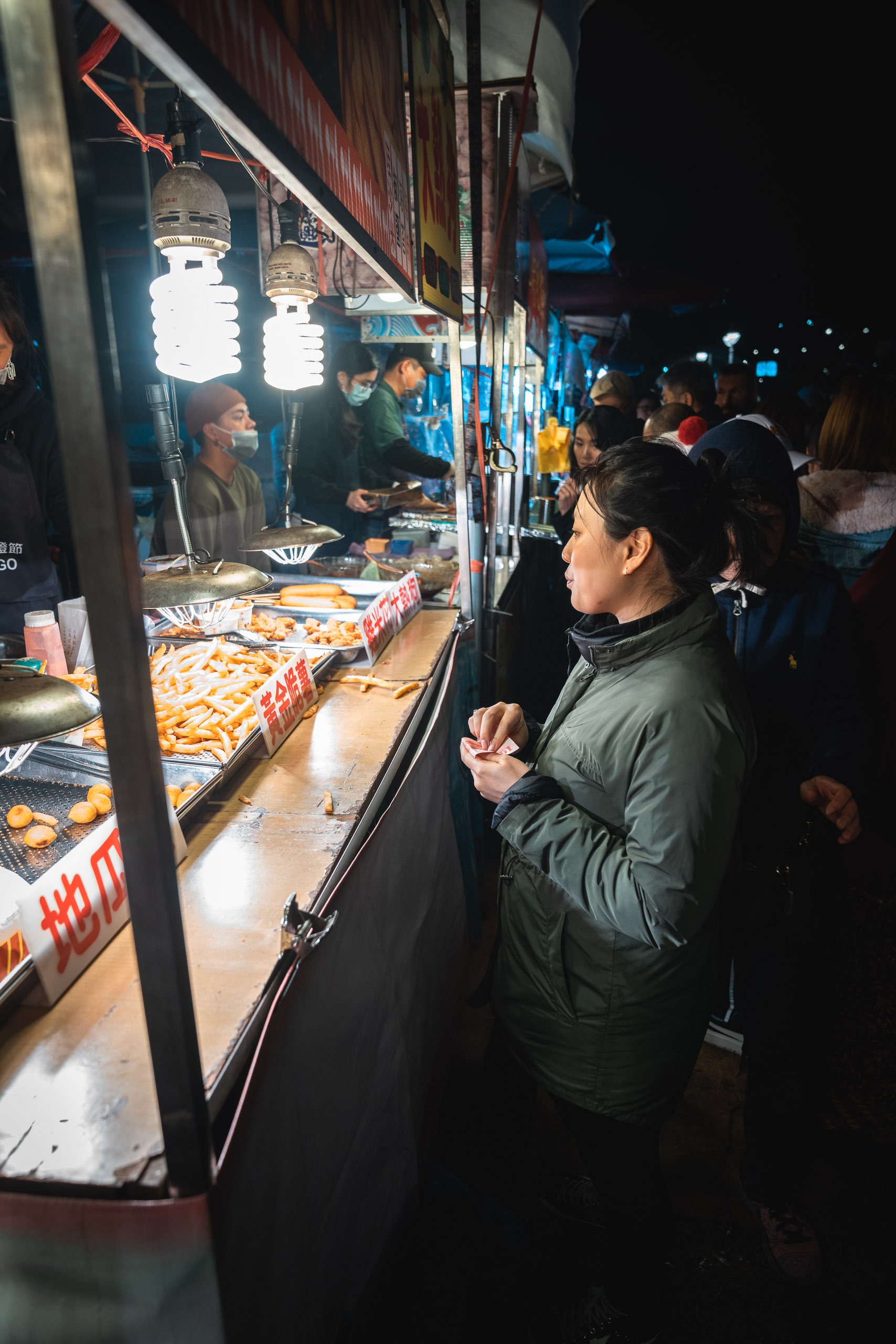 A customer in a dark jacket waits at a brightly lit nighttime food stall displaying golden fried items, with vendors inside and a crowd of people gathered along the street