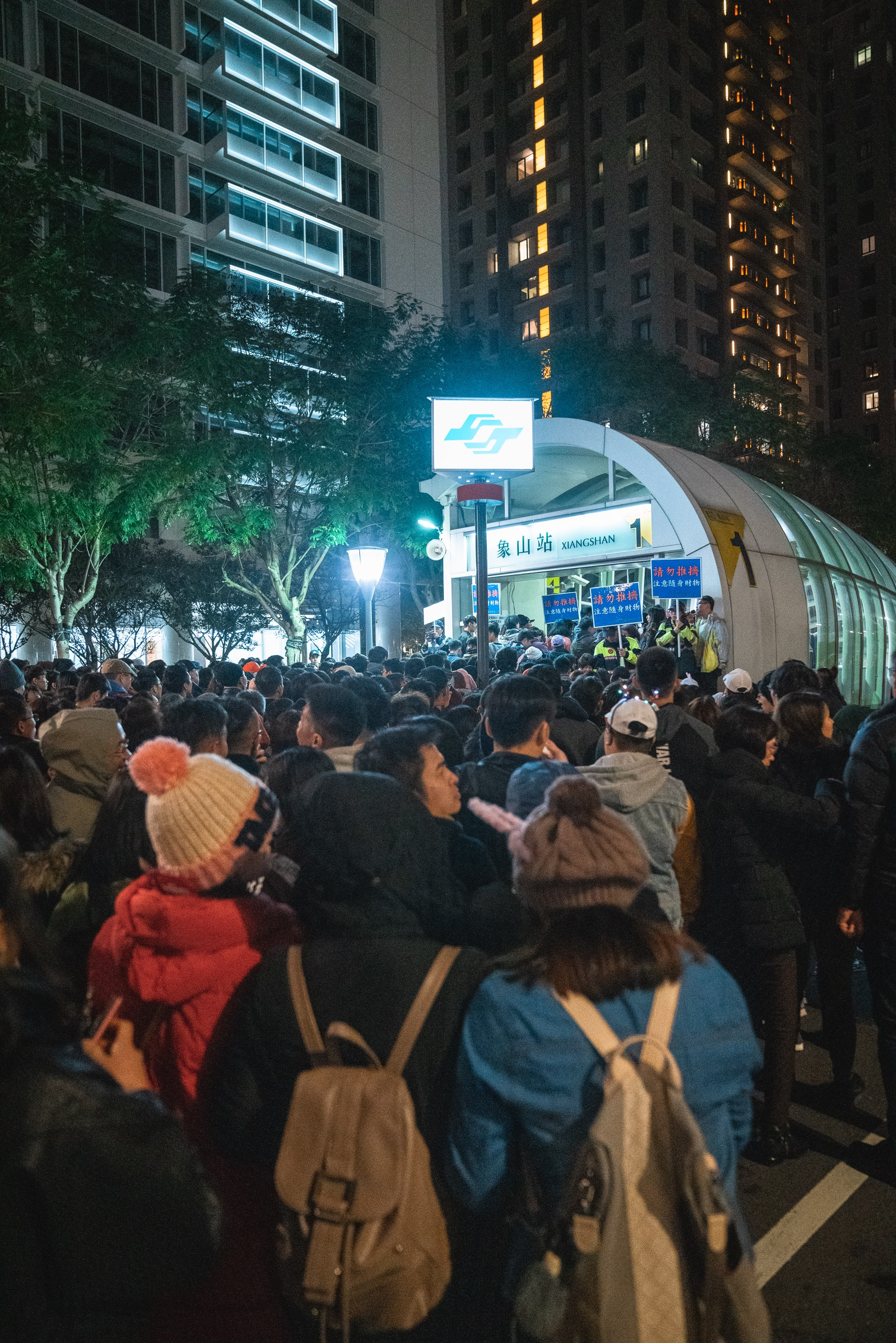 Dense crowds of commuters gather at an illuminated subway station entrance at night, surrounded by glowing high-rise apartment buildings and bare trees in urban Taiwan