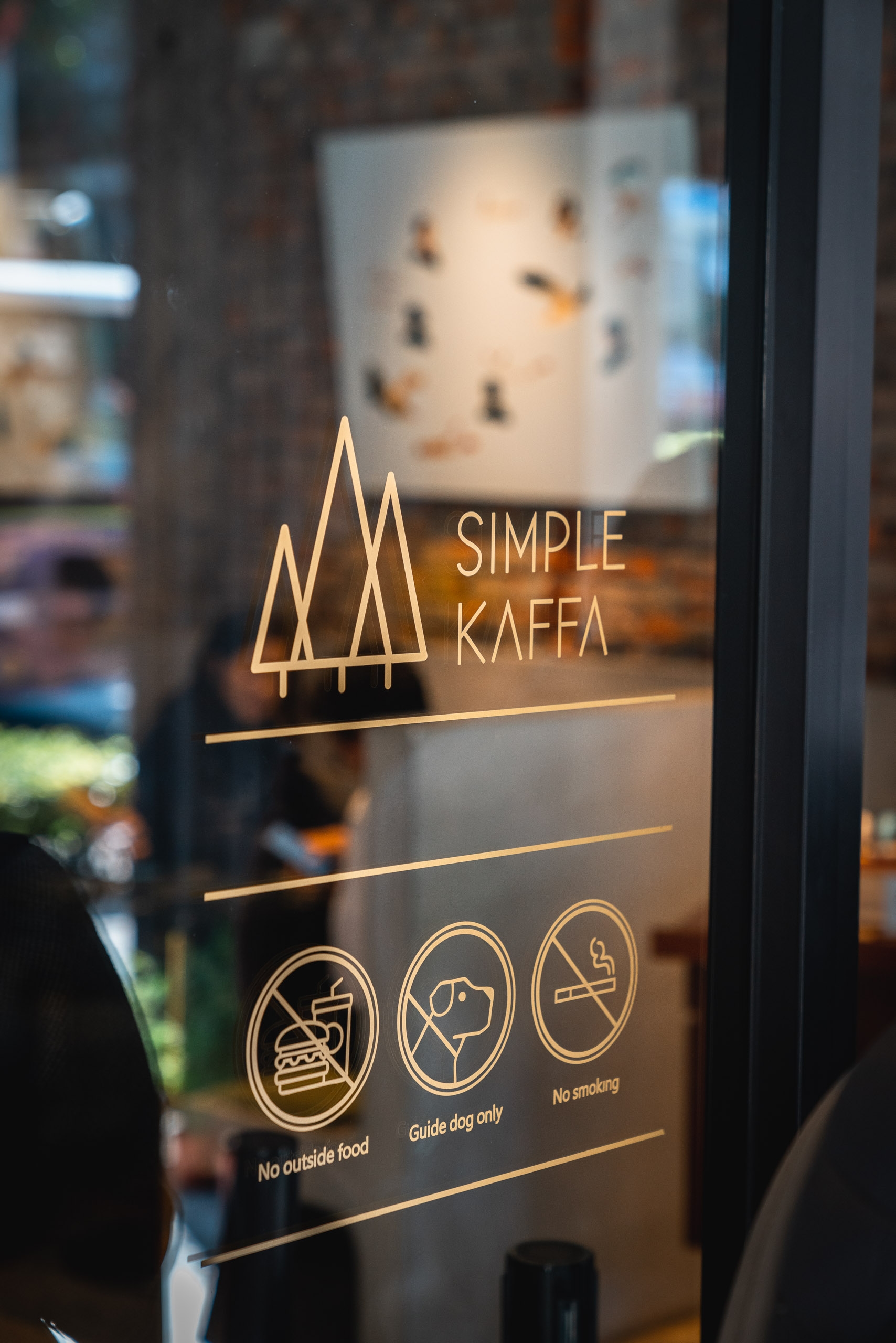 Gold etched signage for "Simple Kaffa" coffee shop displays on a glass door with icons indicating house rules about outside food, guide dogs, and no smoking policies