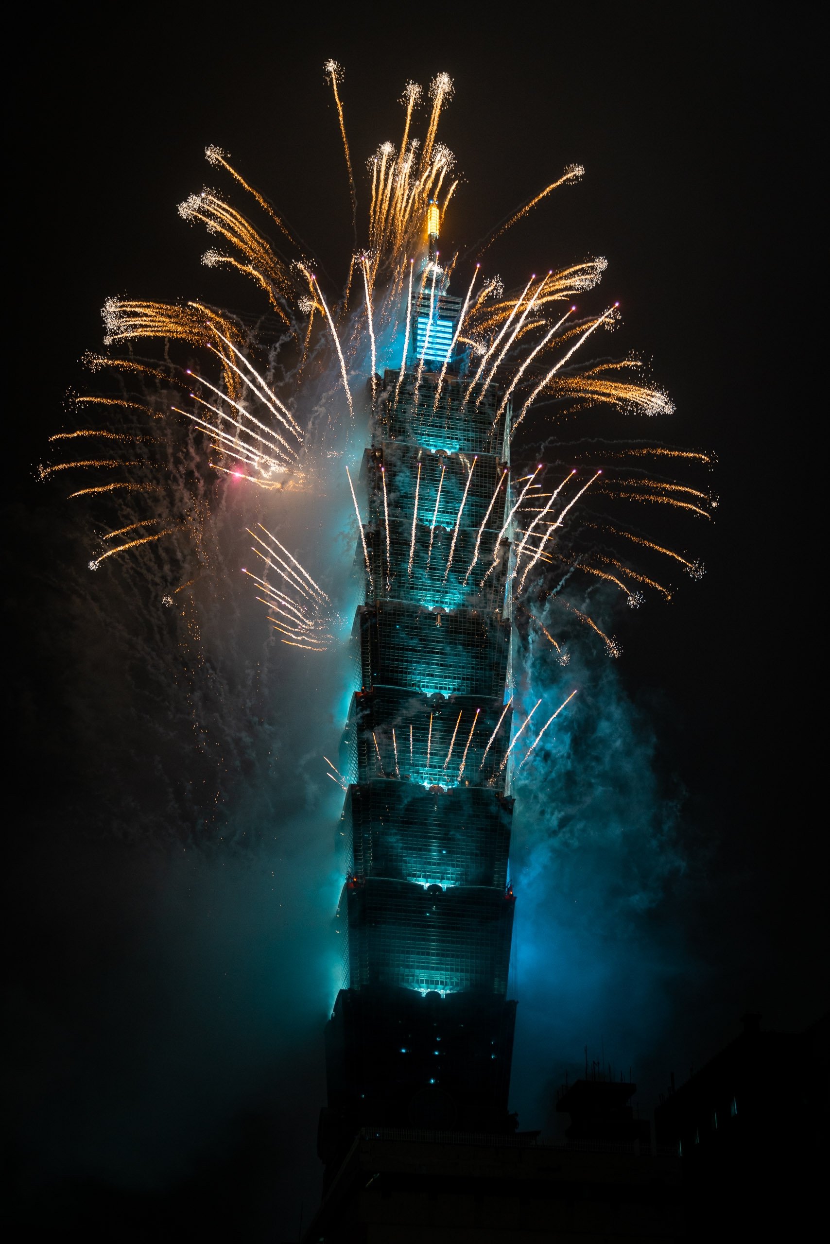 A towering skyscraper illuminated in cyan blue light erupts with golden fireworks bursting outward against the dark night sky, with smoke swirling around its illuminated facade