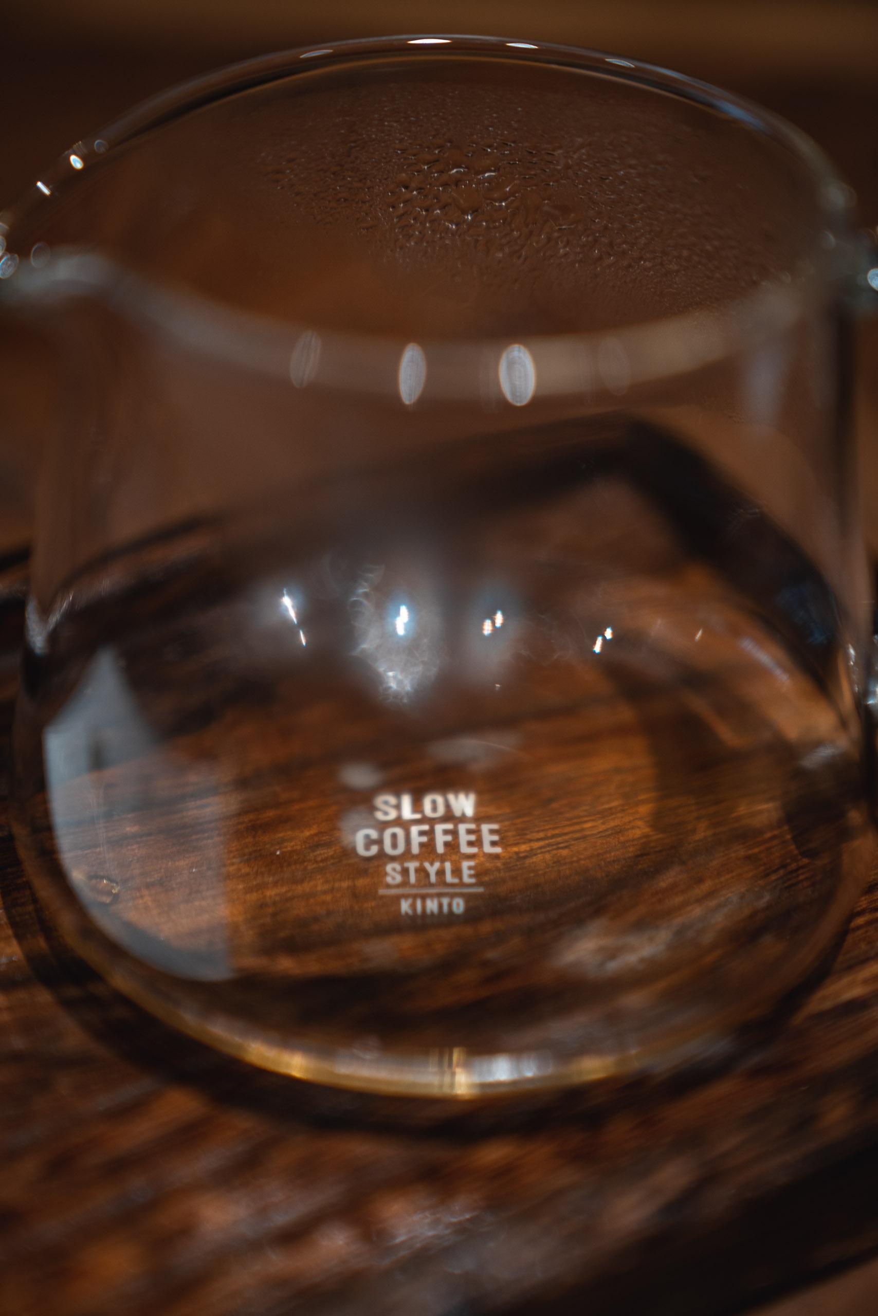 A clear glass carafe labeled "Slow Coffee Style Kinto" sits on a wooden surface, filled with dark brewed coffee under warm indoor lighting