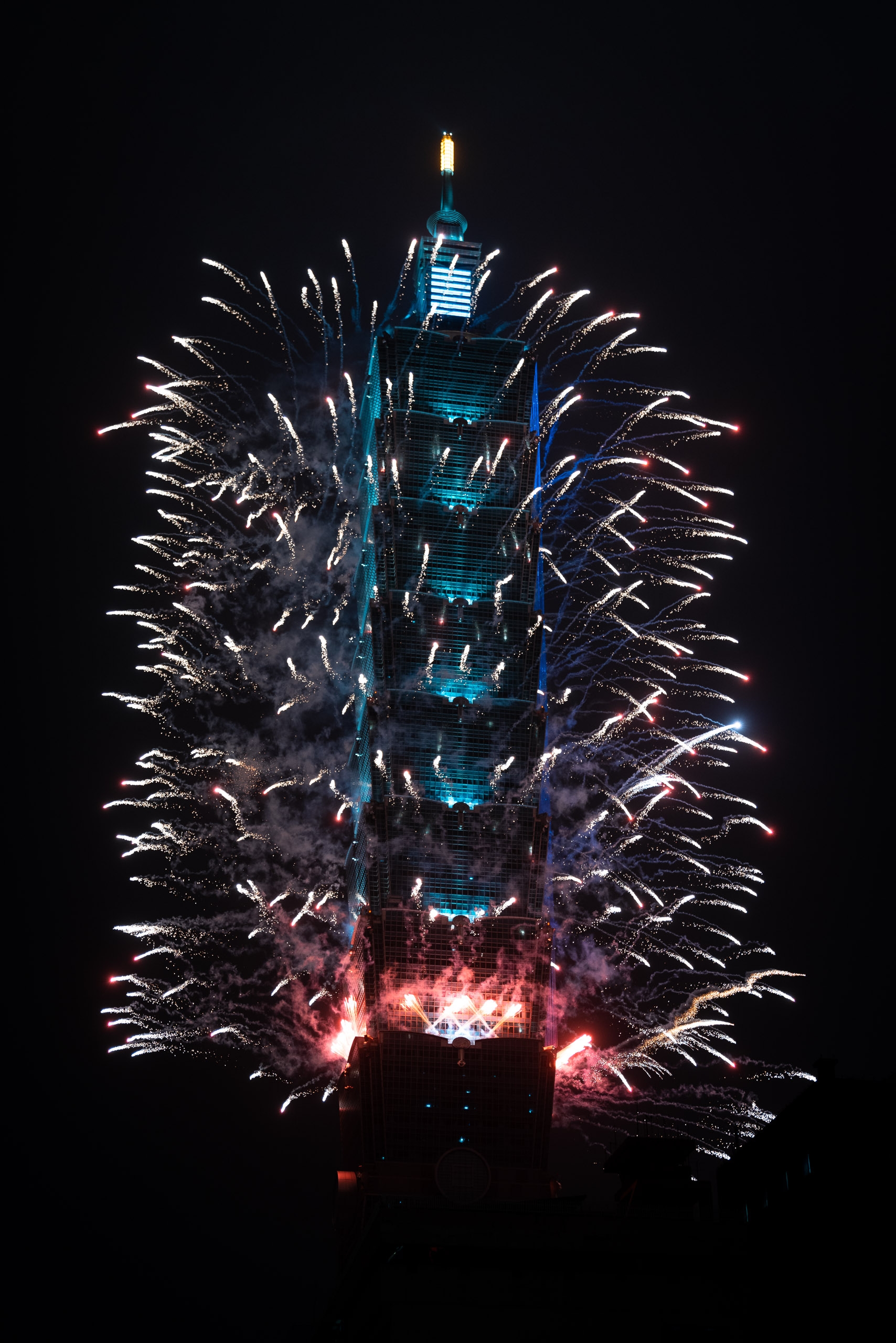 Taipei 101 tower illuminated in blue and gold stands against the night sky while symmetrical fireworks burst in white and red streaks on both sides