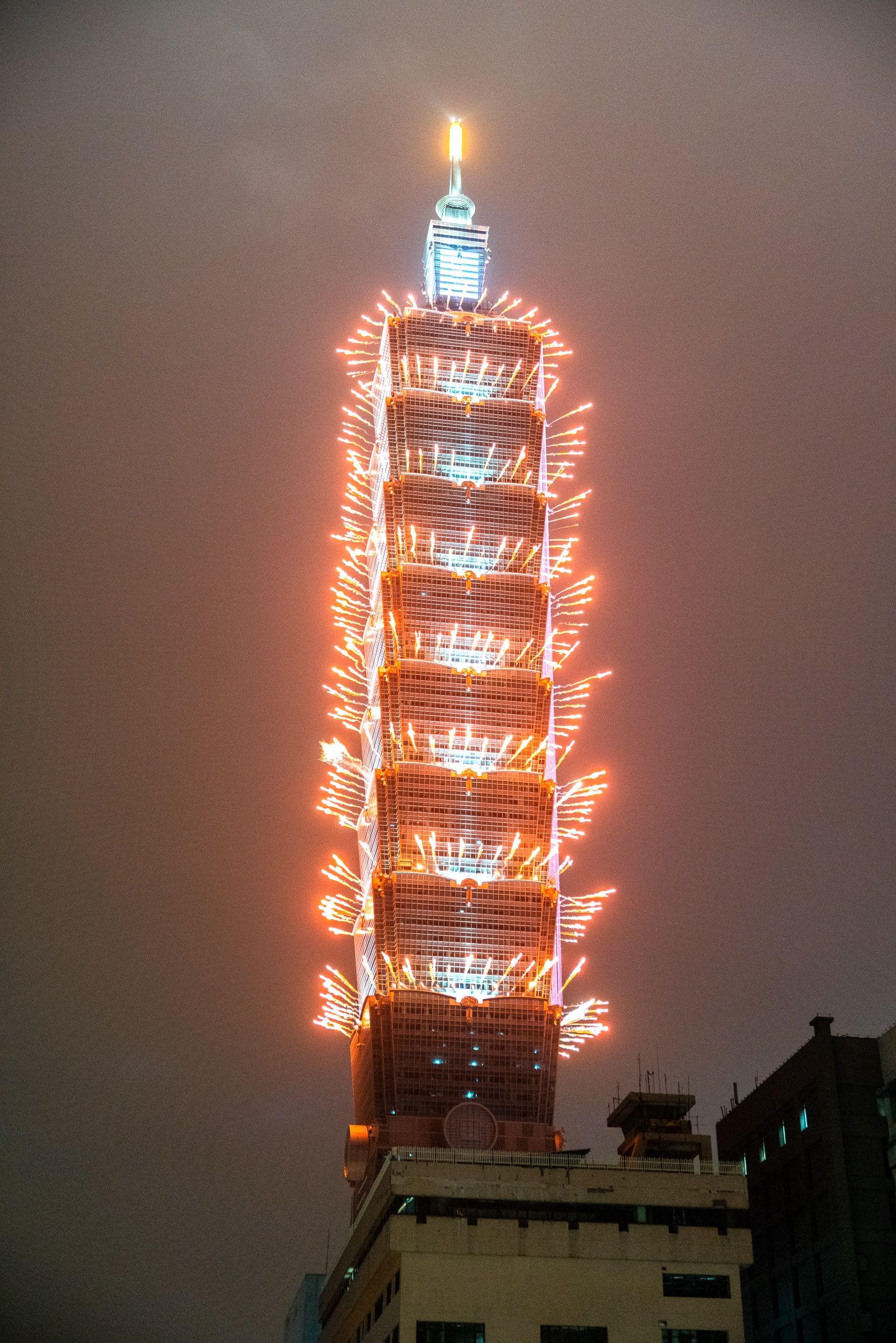 Taipei 101's illuminated facade glows with warm orange neon lights outlining each floor against a hazy twilight sky, with a bright beacon crowning its spire